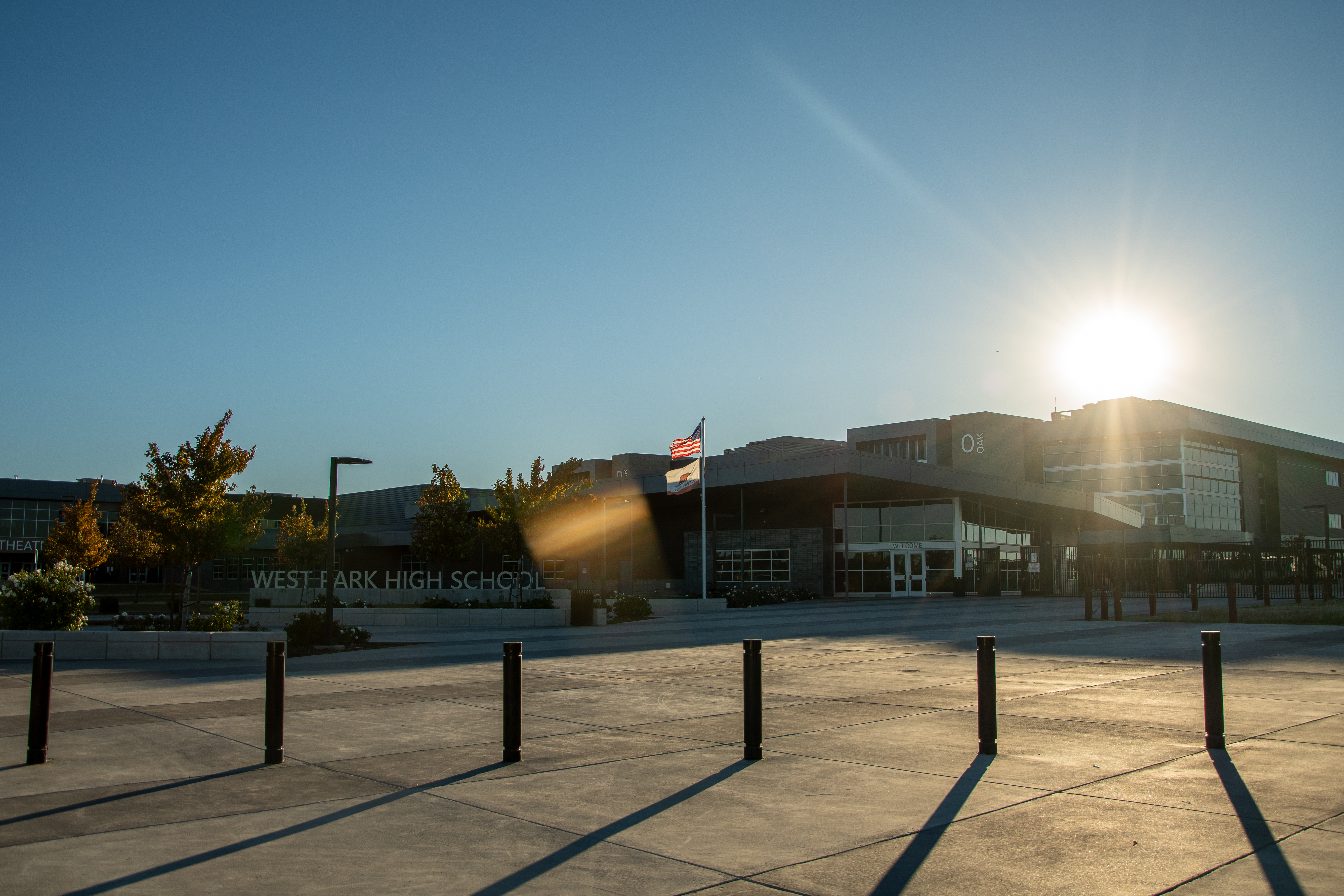 A sunny outdoor school campus with a concrete plaza in the foreground and a high school building in the background, surrounded by trees and a clear blue sky.