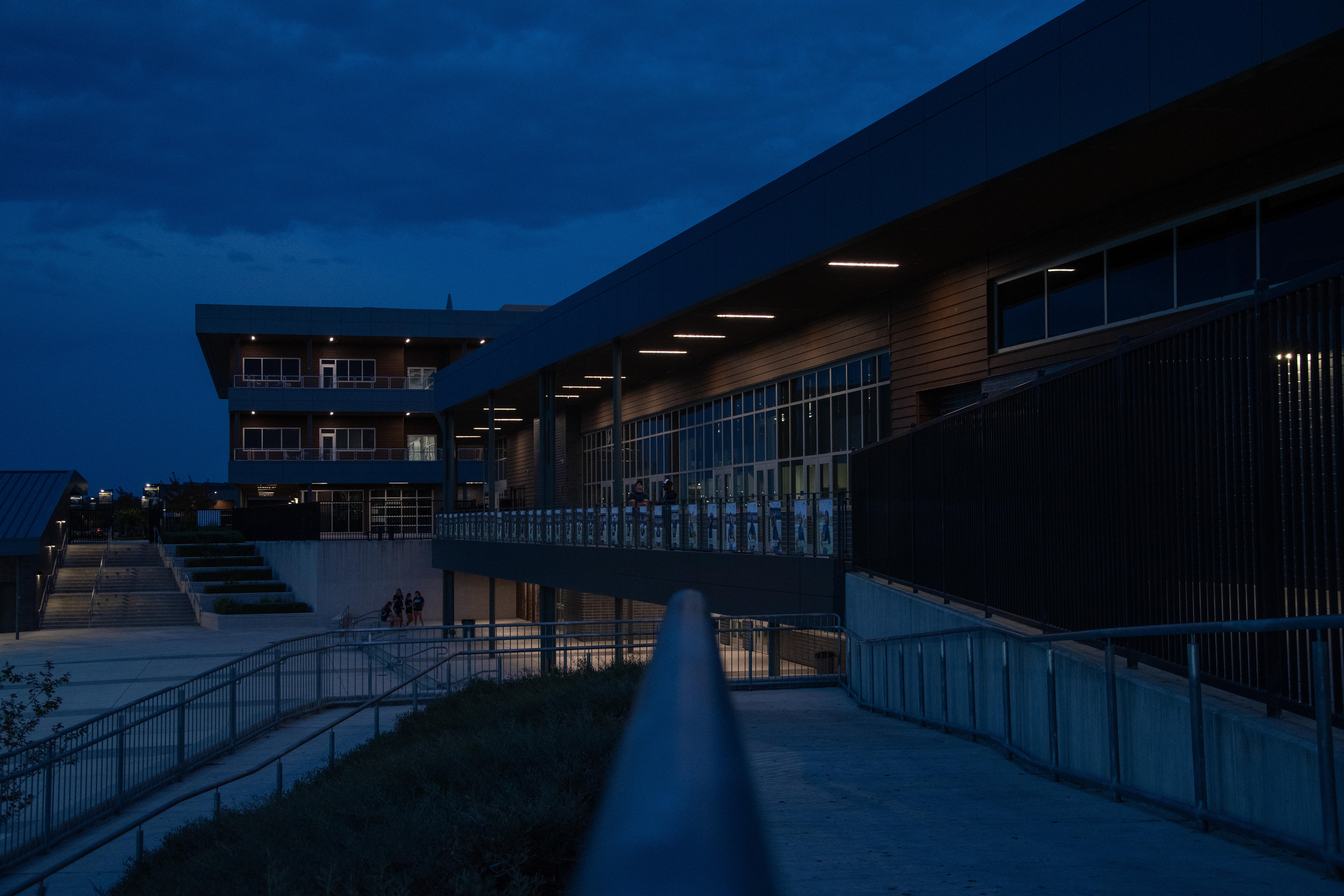 A dimly lit walkway leads towards a large, multi-story building with illuminated windows against a dark, cloudy sky.