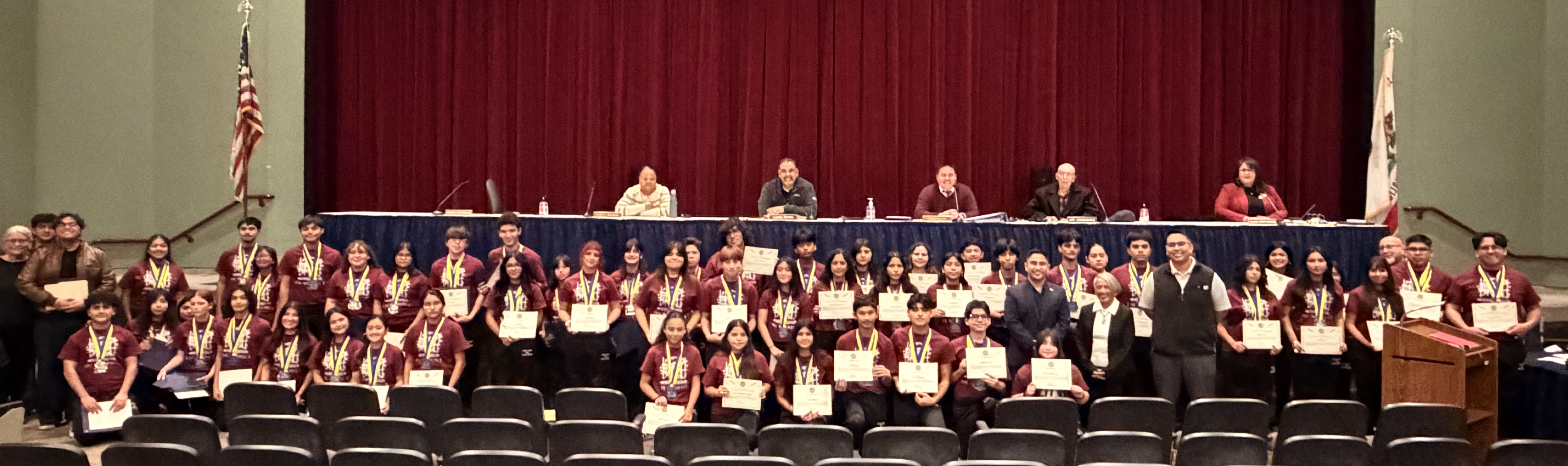 A large group of people, likely students, are seated in rows on a stage with a red curtain backdrop, some wearing ceremonial robes and holding certificates or diplomas.