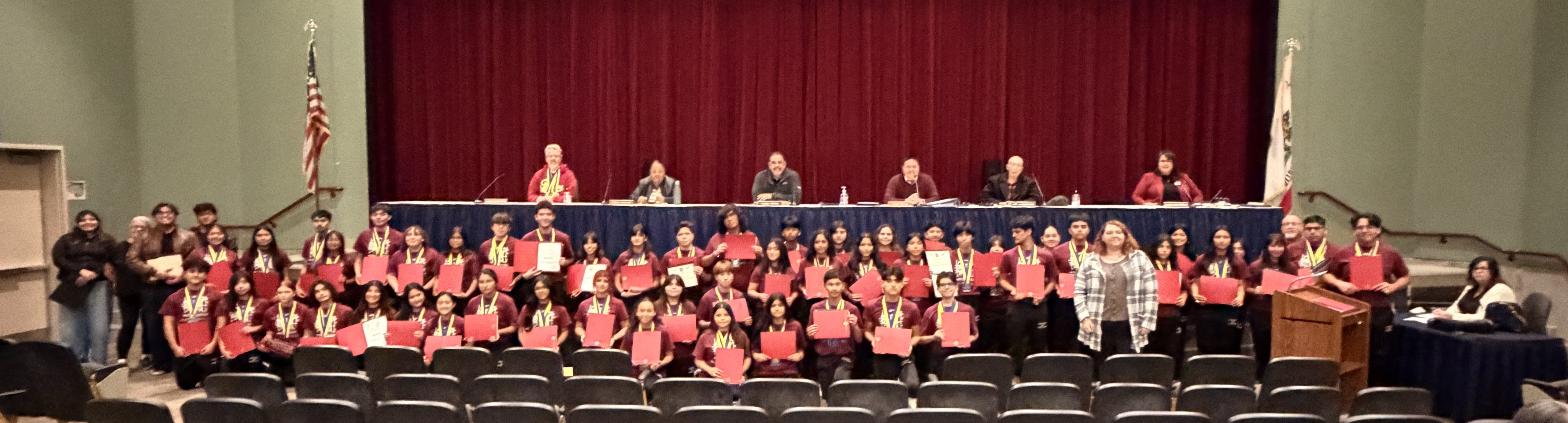 A stage with a red curtain backdrop, featuring a panel of people seated at a table in the foreground and a large audience in the background wearing red shirts.