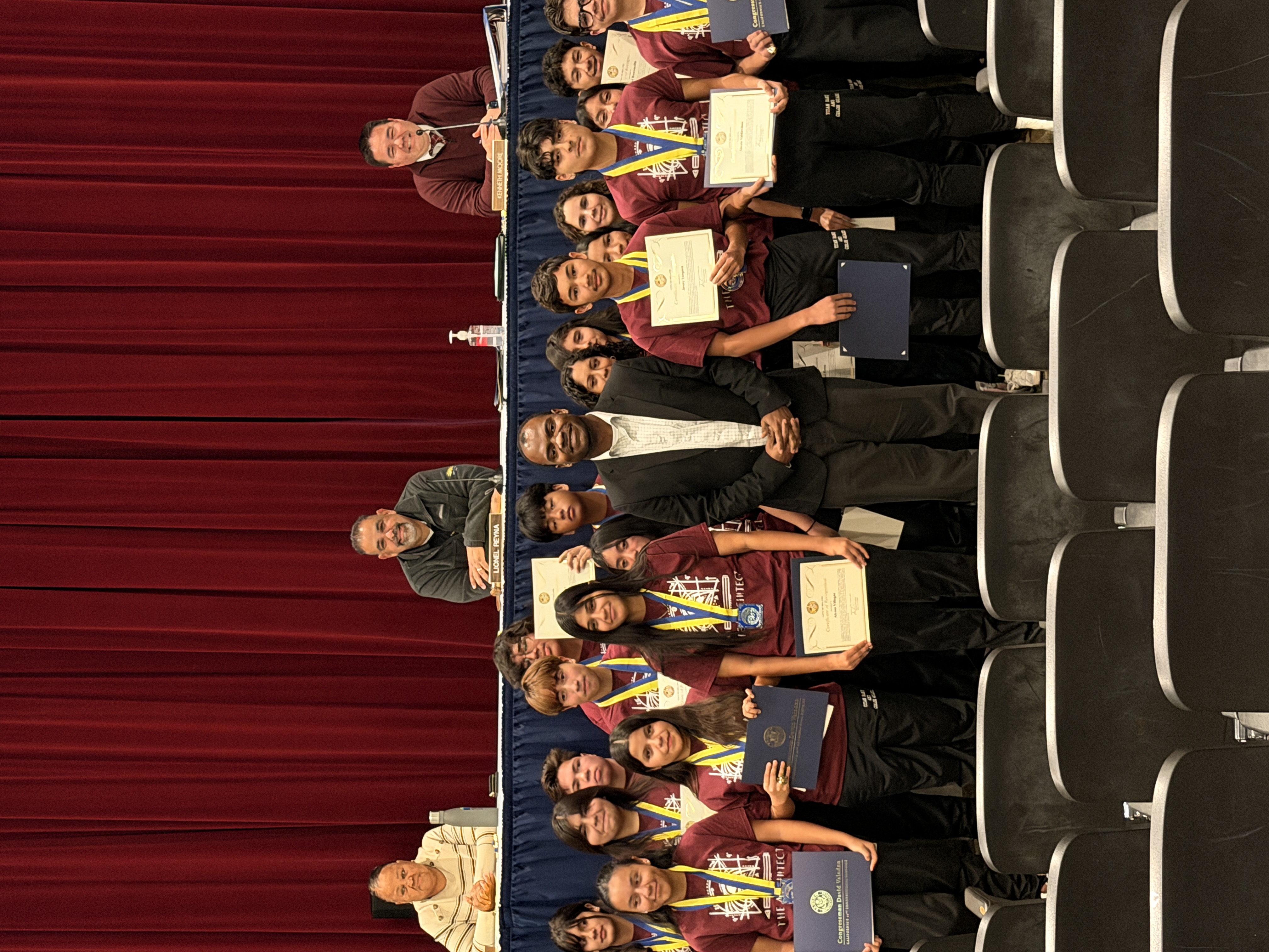 A group of young students, some holding certificates, are seated in front of a panel of adults on a stage with a red curtain backdrop.