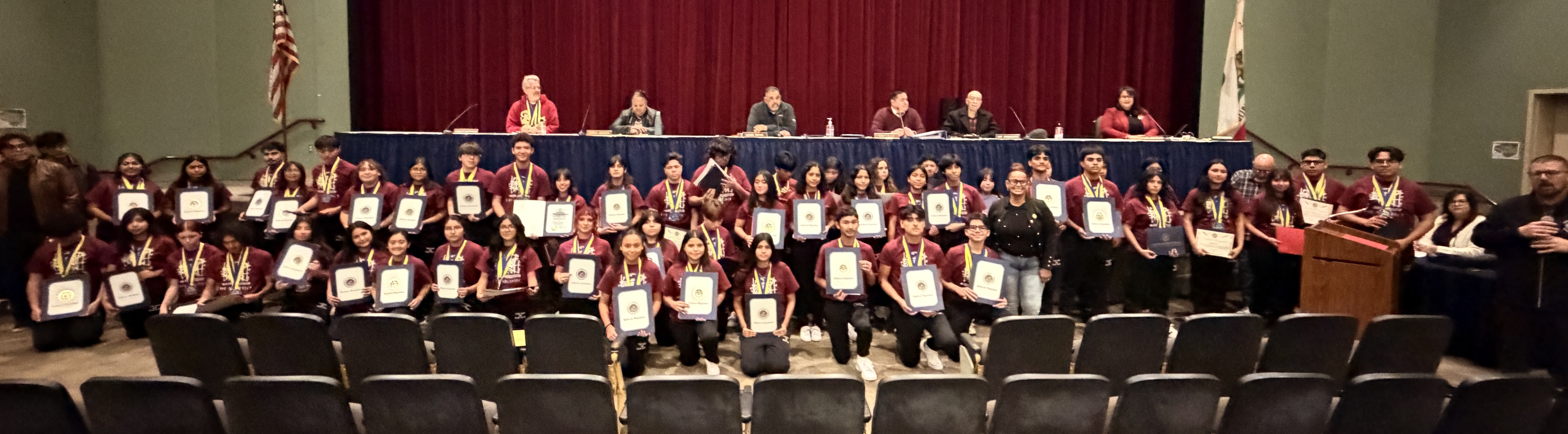 A large group of people, likely students, are seated in rows on stage, holding certificates or awards, with a panel of adults seated behind them on a raised platform.
