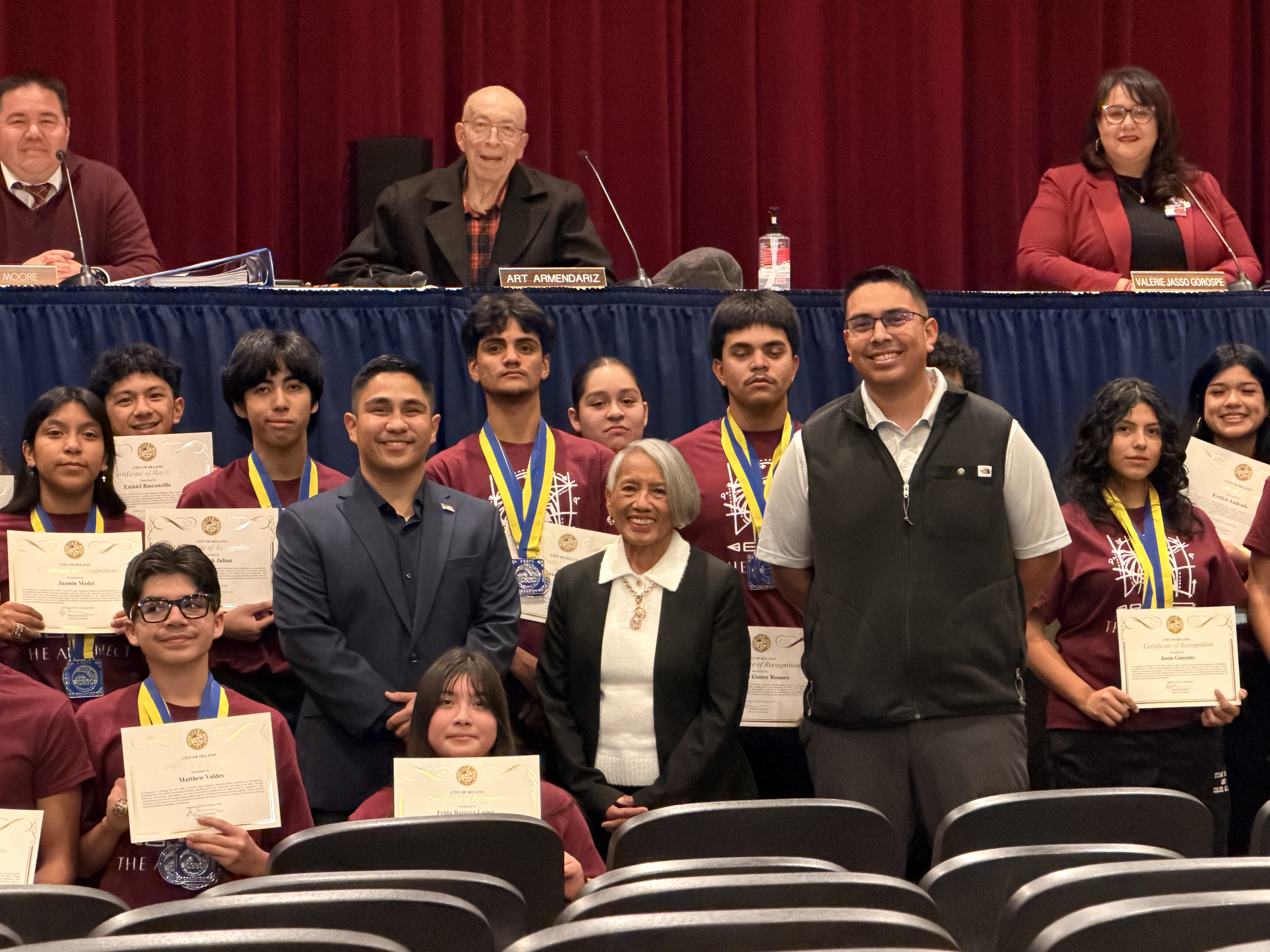 The image shows a group of students and faculty members gathered on a stage, with a red curtain backdrop. The students are holding certificates and appear to be celebrating an achievement or graduation.