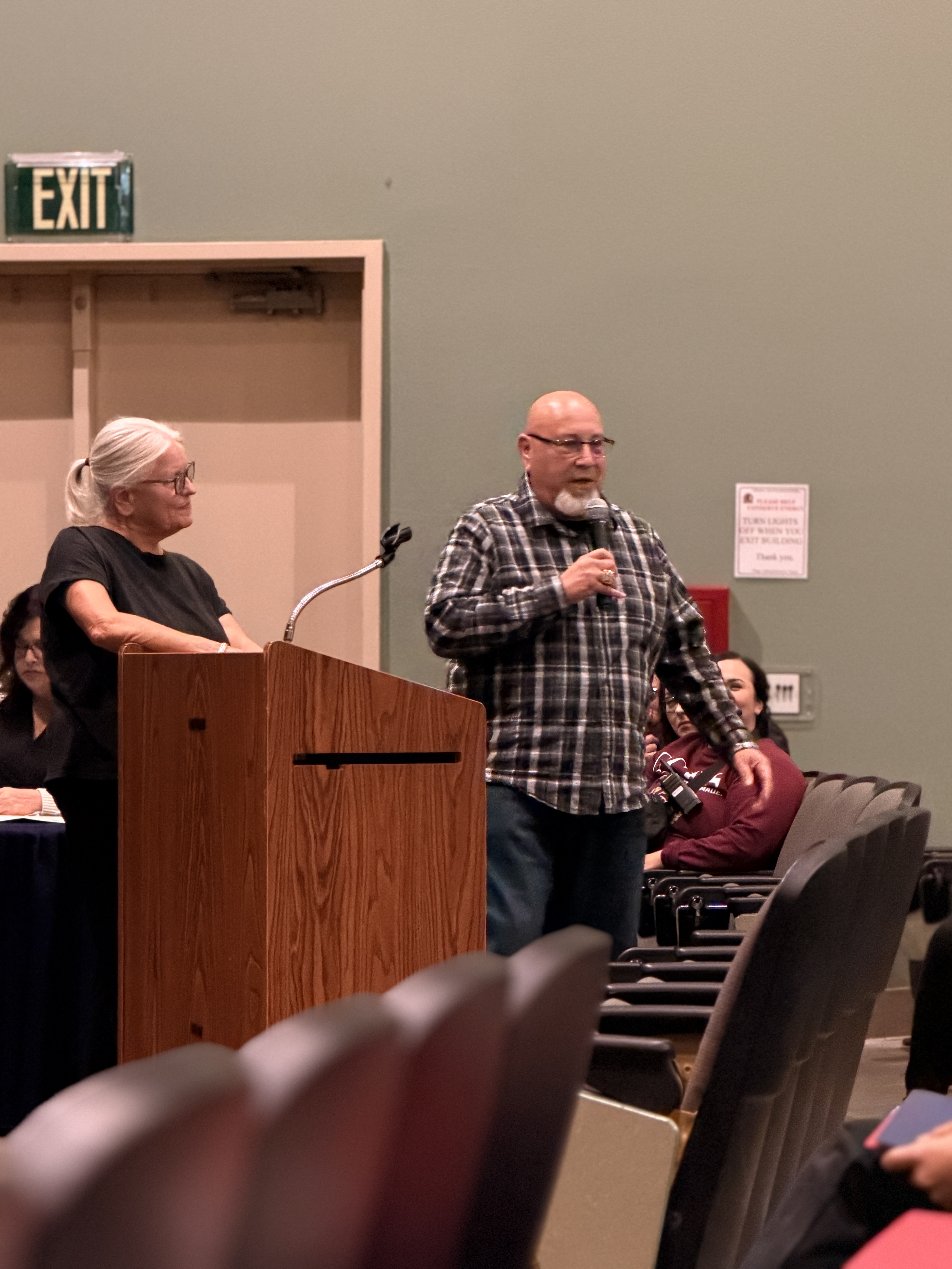 The image shows two people standing at a podium in what appears to be a meeting or conference room, with an "EXIT" sign visible in the background.