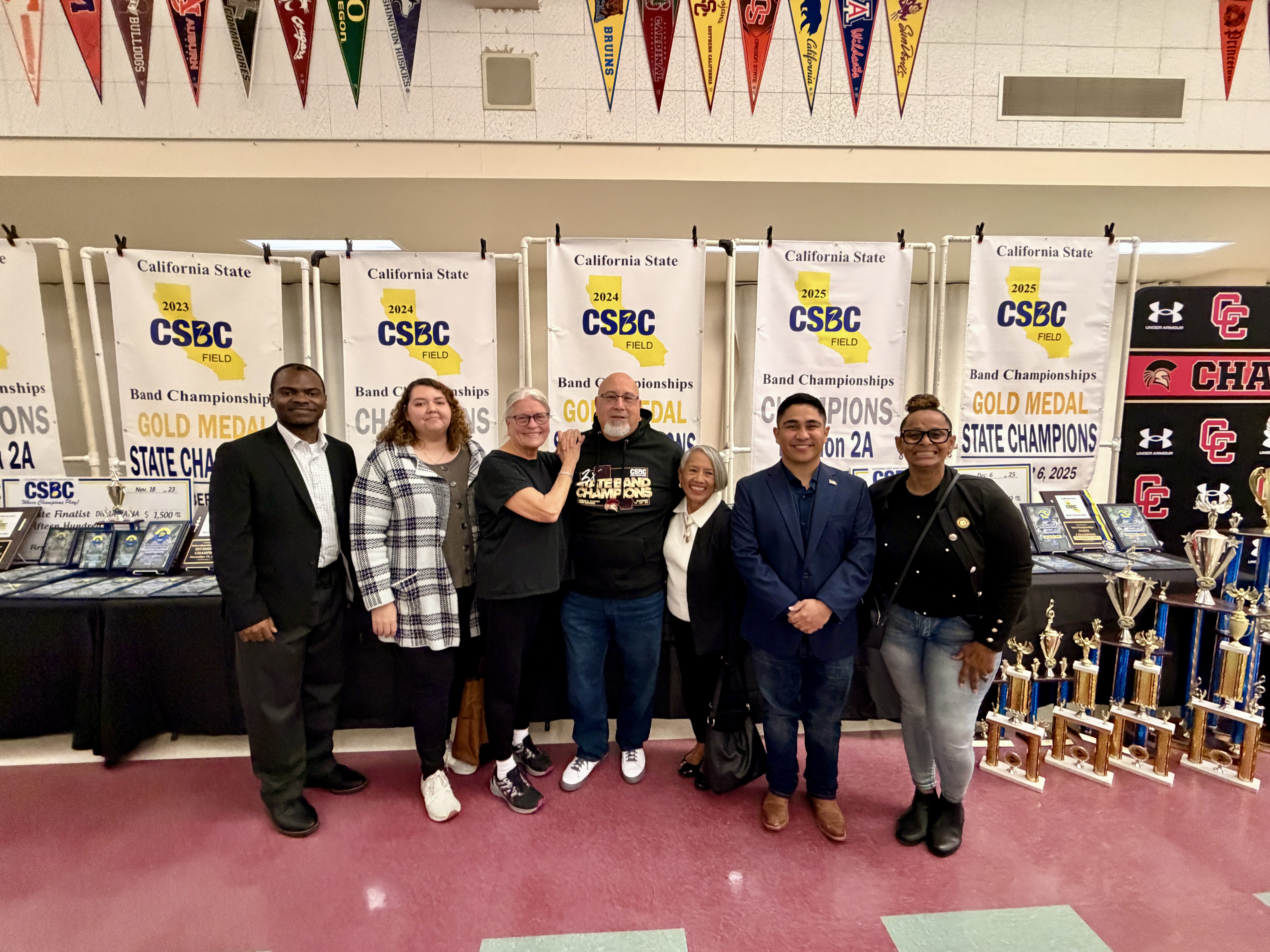 The image shows a group of people standing in front of a display of sports championship banners and trophies.