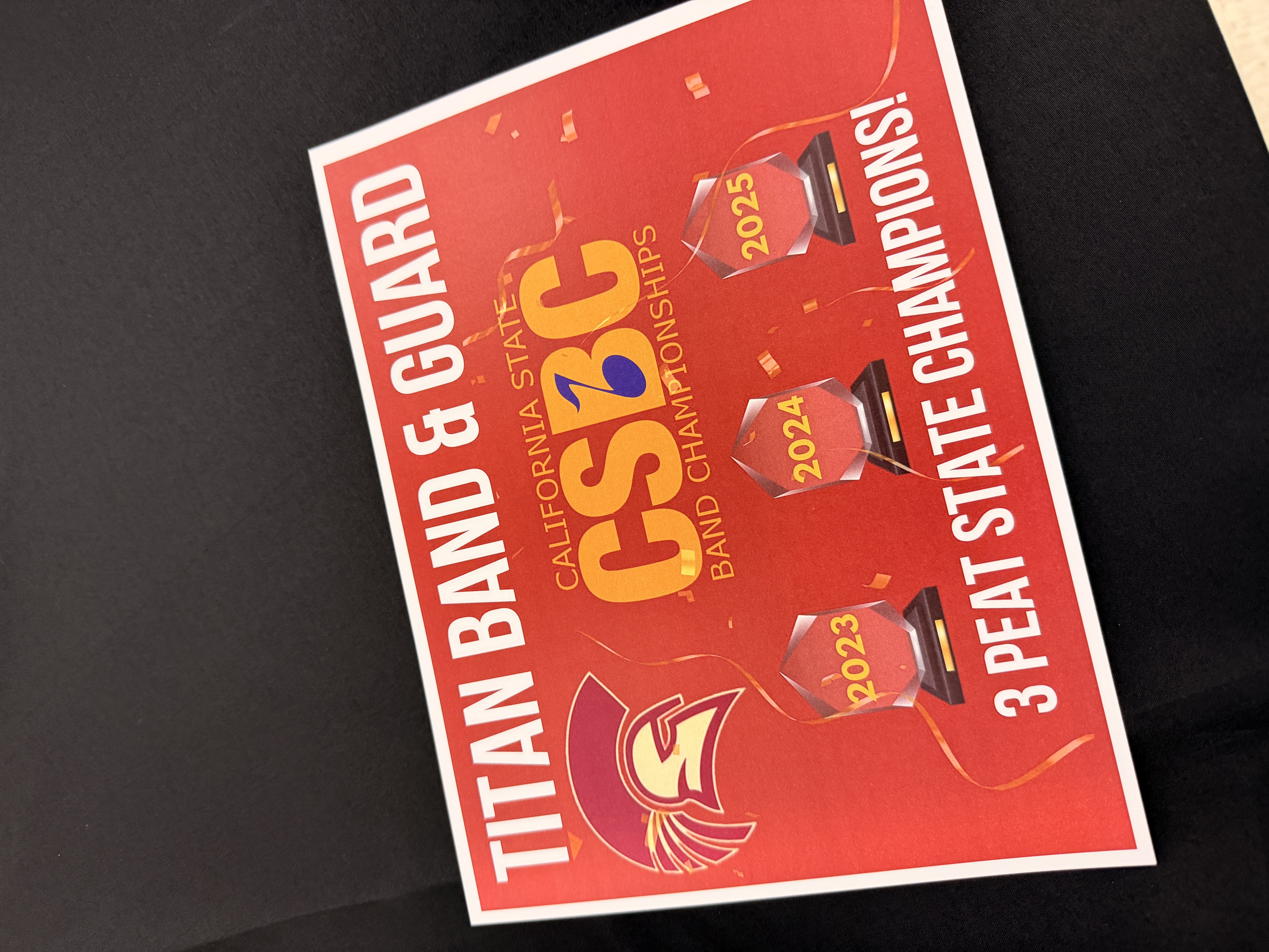 The image shows a red and yellow banner with the text "TITAN BAND &amp; GUARD" and "CSB BAND CHAMPIONSHIPS" along with the years 2023, 2024, and 2025, indicating three consecutive state championship wins.