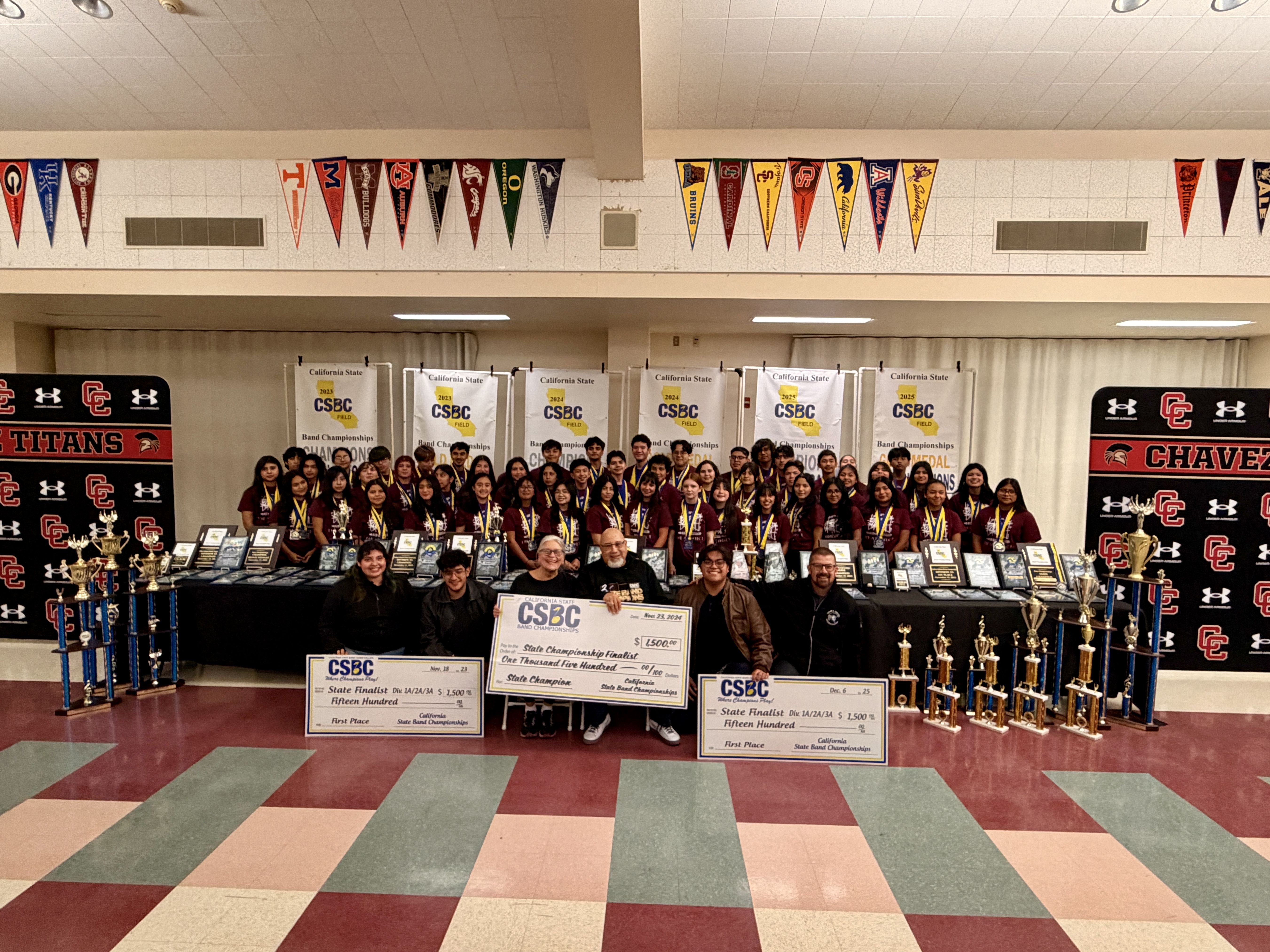 A large group of people, likely students, posing for a photograph in a school setting, surrounded by sports equipment, trophies, and banners.