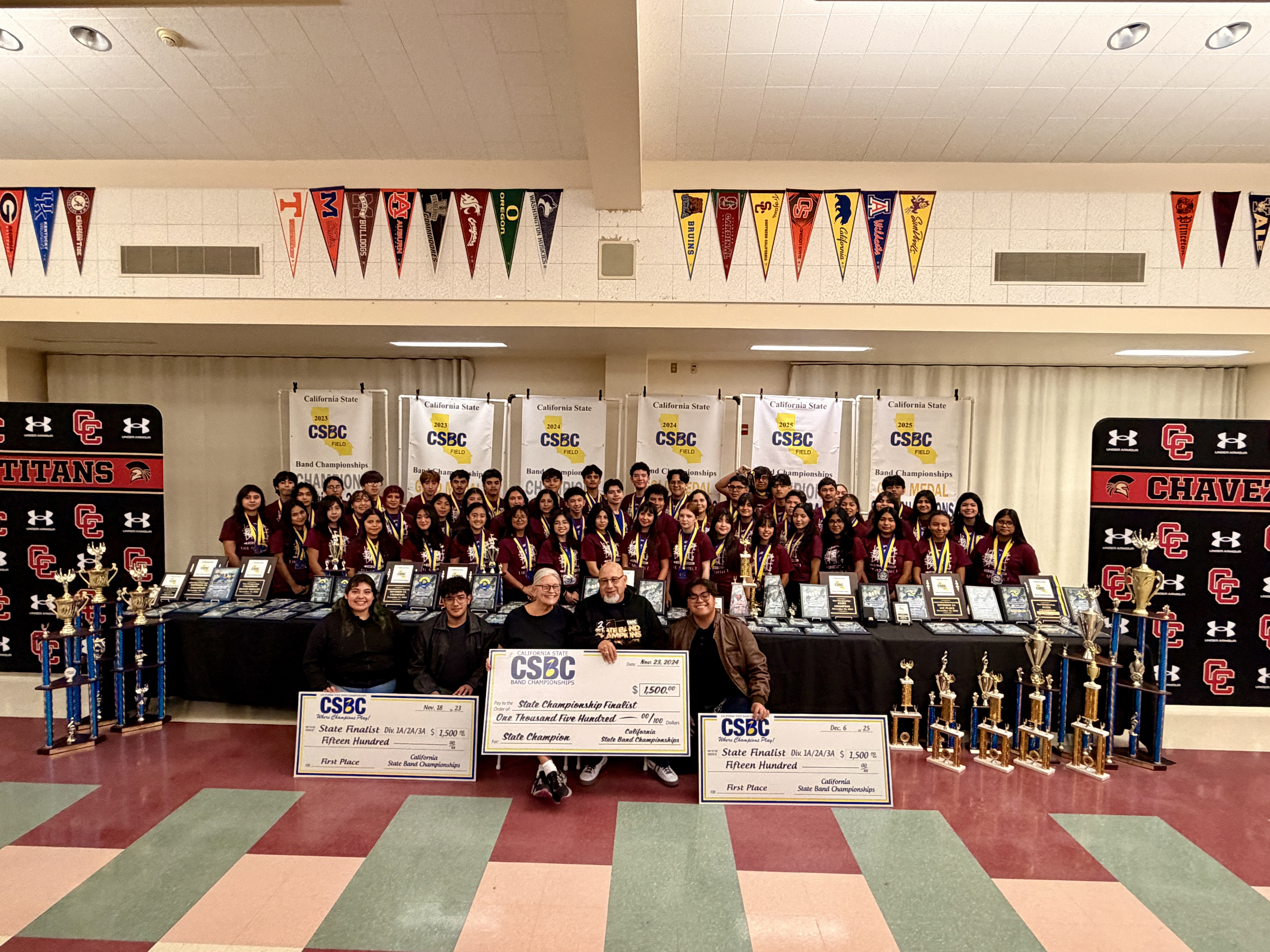 A large group of people, likely students, posing together in a school setting, surrounded by sports equipment, trophies, and school banners.