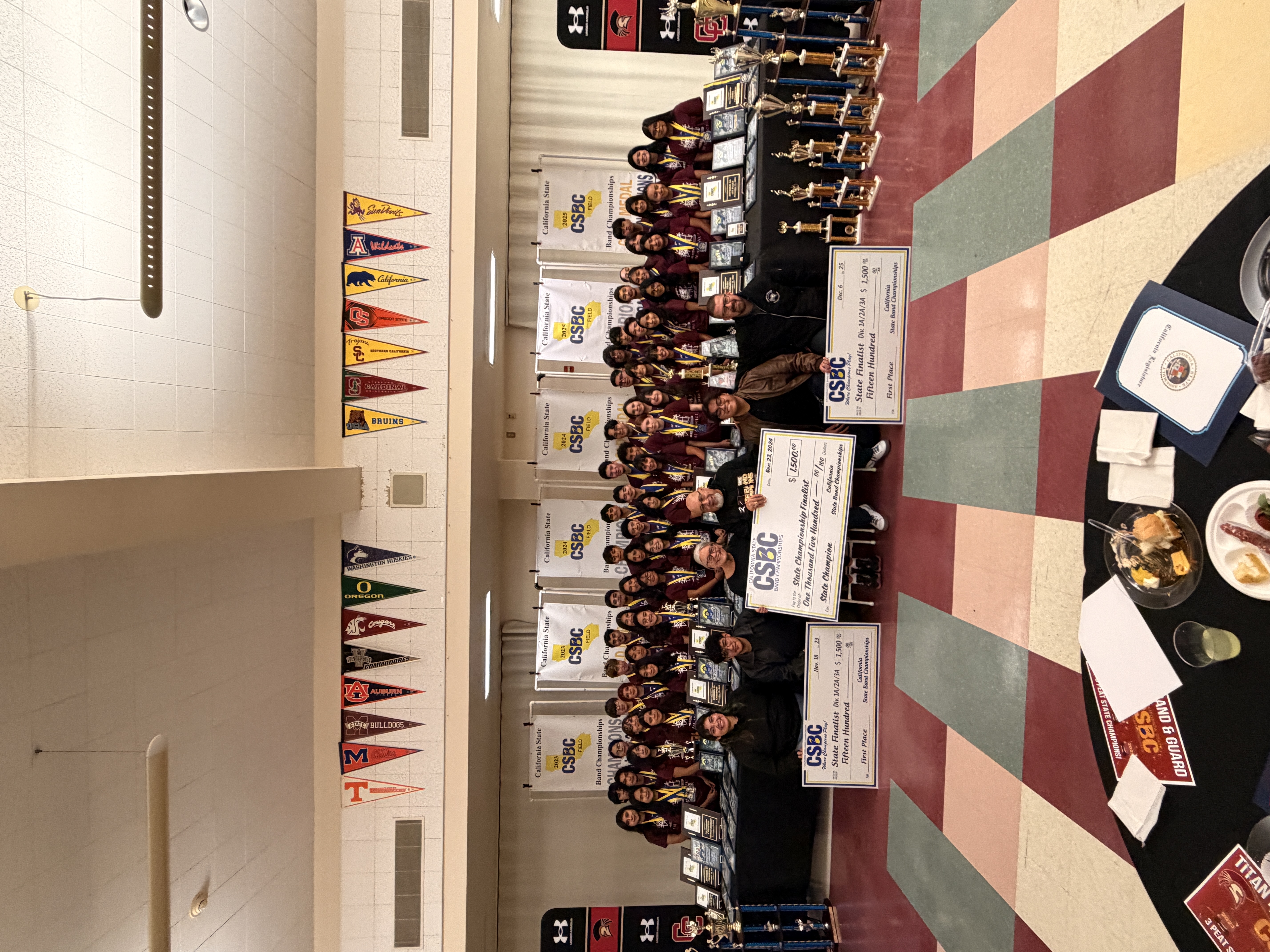 A large group of people, likely a sports team or club, posing together in a gymnasium-like setting, with various trophies, banners, and equipment visible in the background.