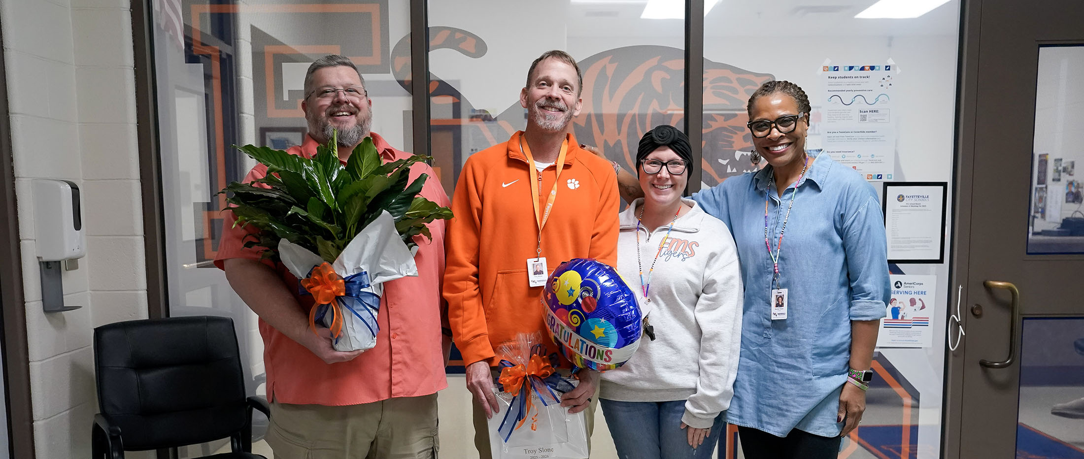 A group of five people, including two men and three women, are standing together in what appears to be an office or workplace setting, with various decorations and artwork visible in the background.