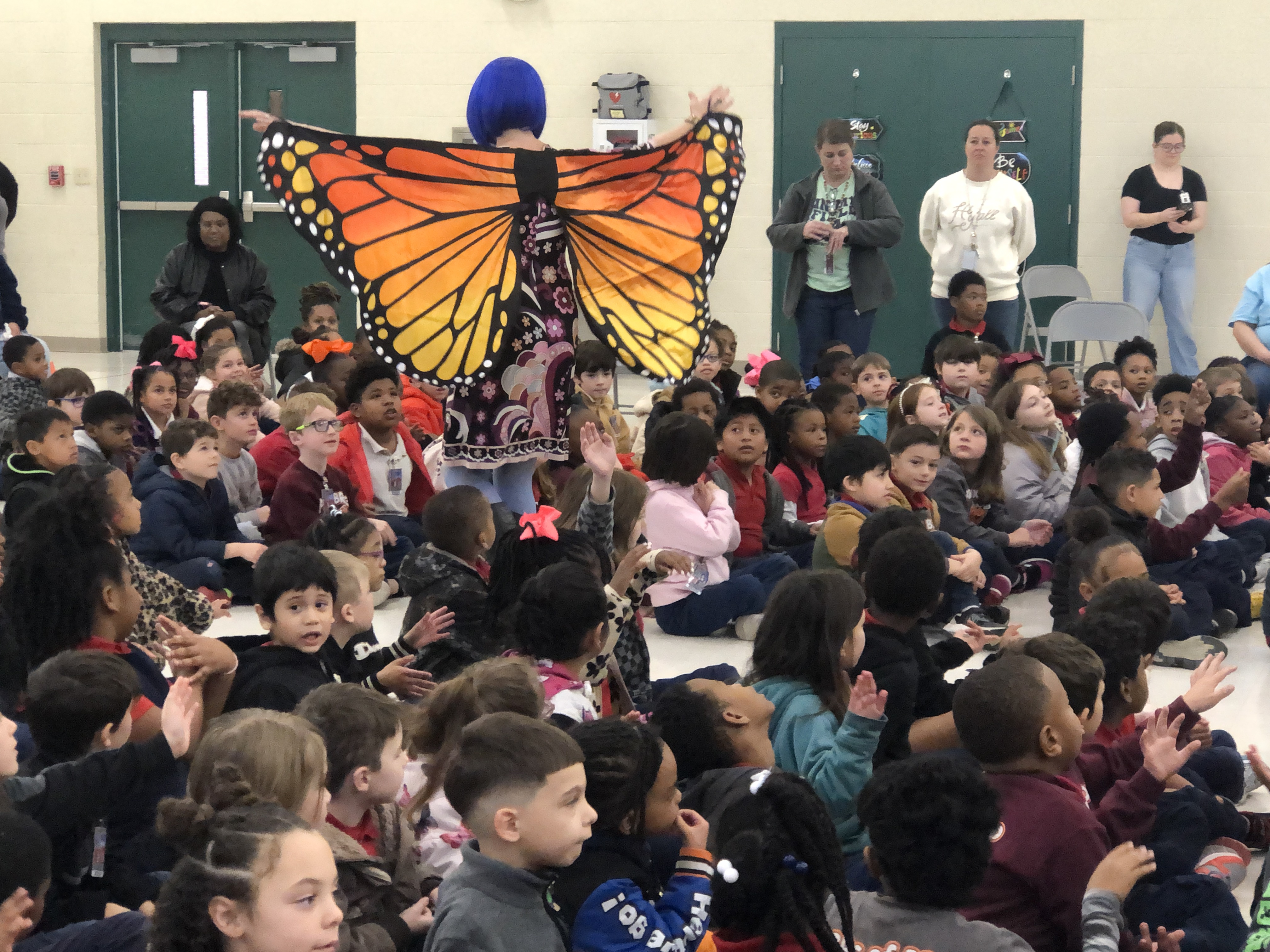 A person in a large butterfly costume stands before a crowd of children sitting on the floor in a gymnasium.