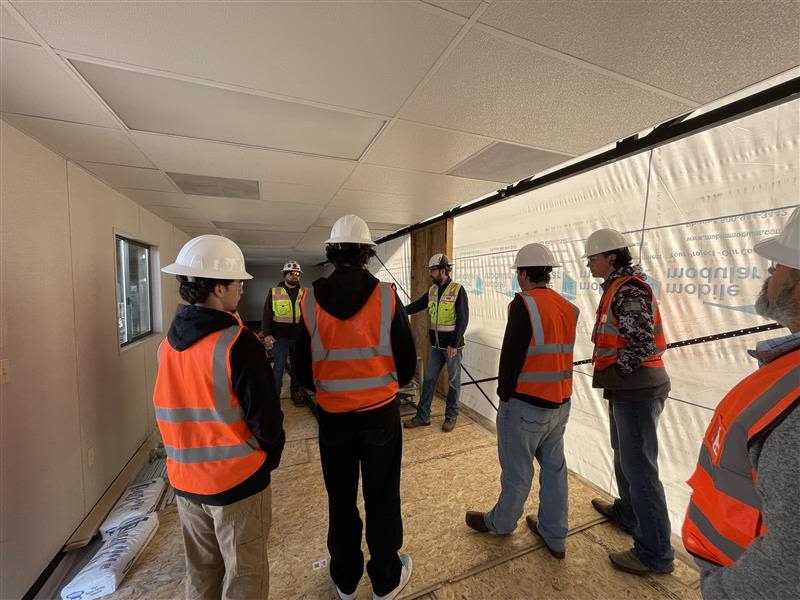 A group of construction workers wearing safety gear, including hard hats and reflective vests, are standing in a hallway of what appears to be an unfinished building or construction site.