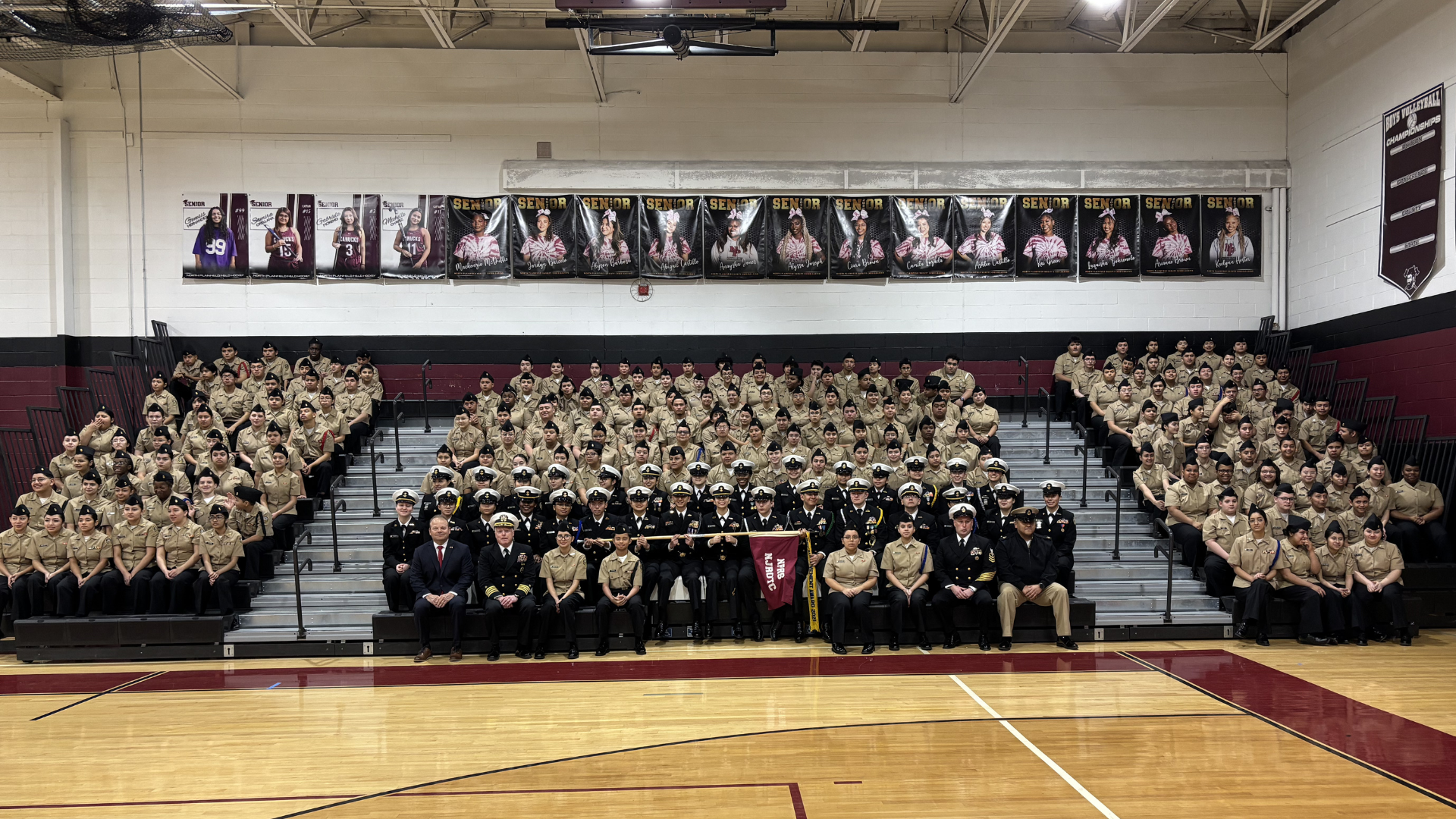 A large group of people, likely a school or organization, gathered in a gymnasium setting, with banners and trophies displayed on the walls.
