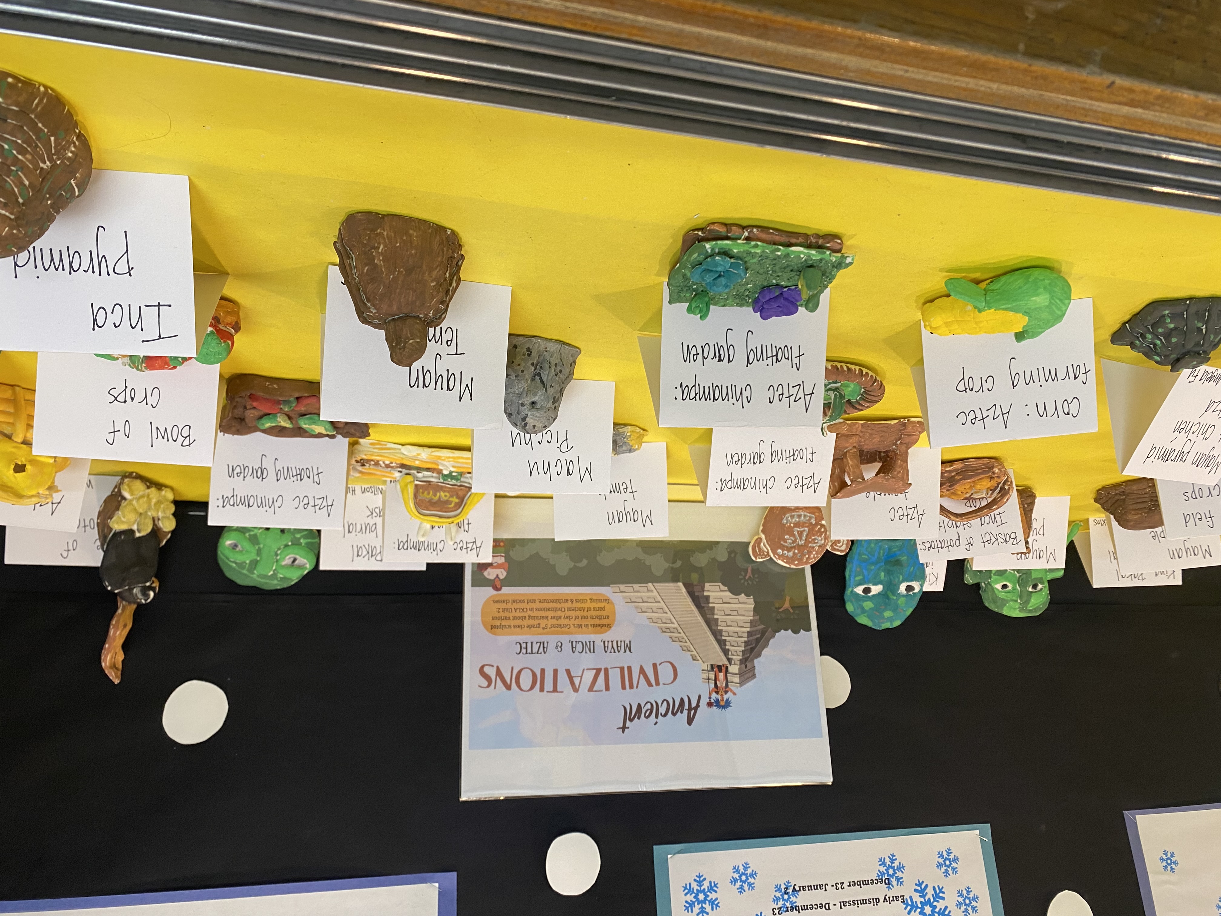 The image shows a display of various models and representations of ancient civilizations, including pyramids, stone structures, and colorful figurines, arranged on a yellow surface with informative labels.