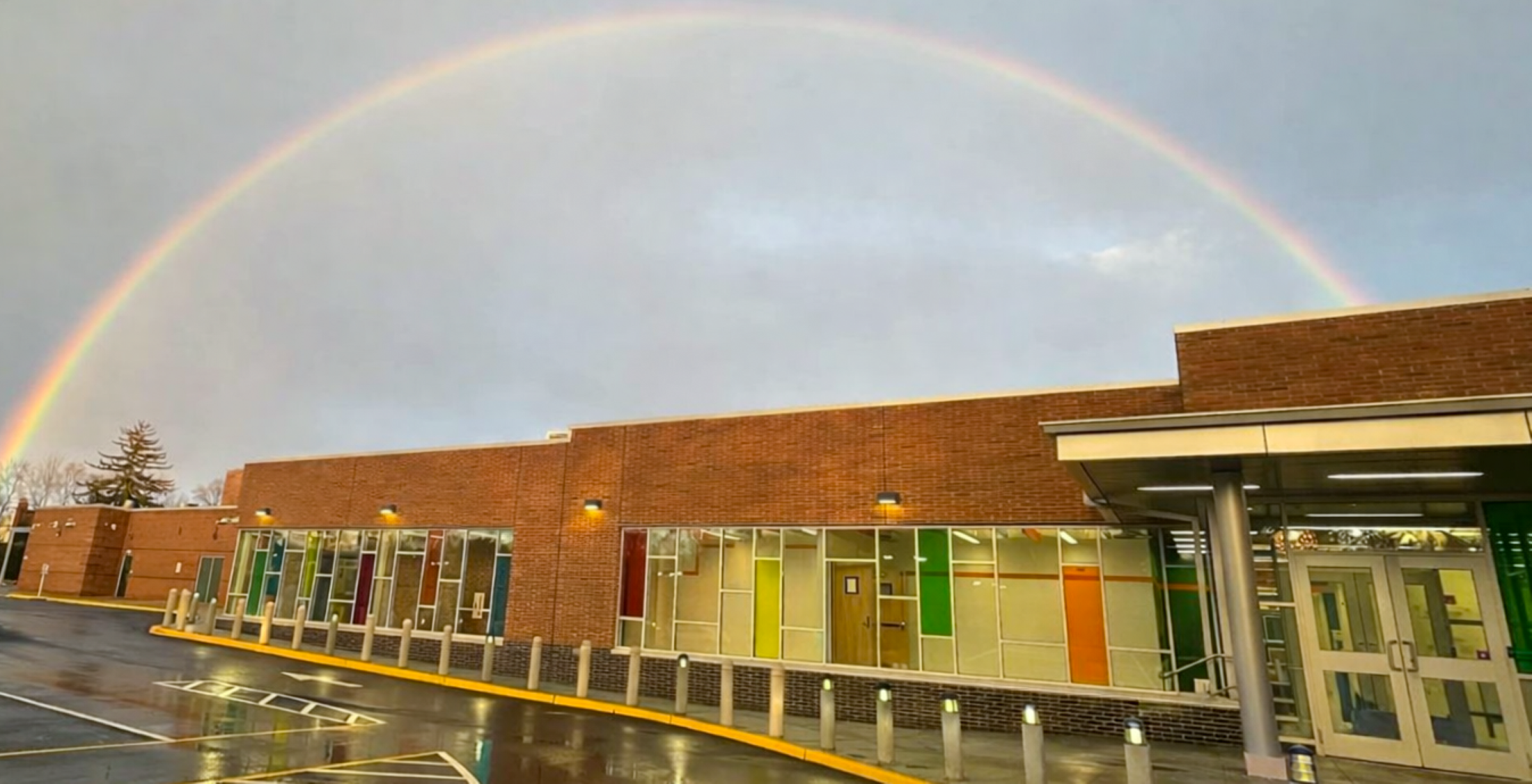 A vibrant rainbow arches across the sky, framing a modern brick building with colorful glass windows in the foreground.