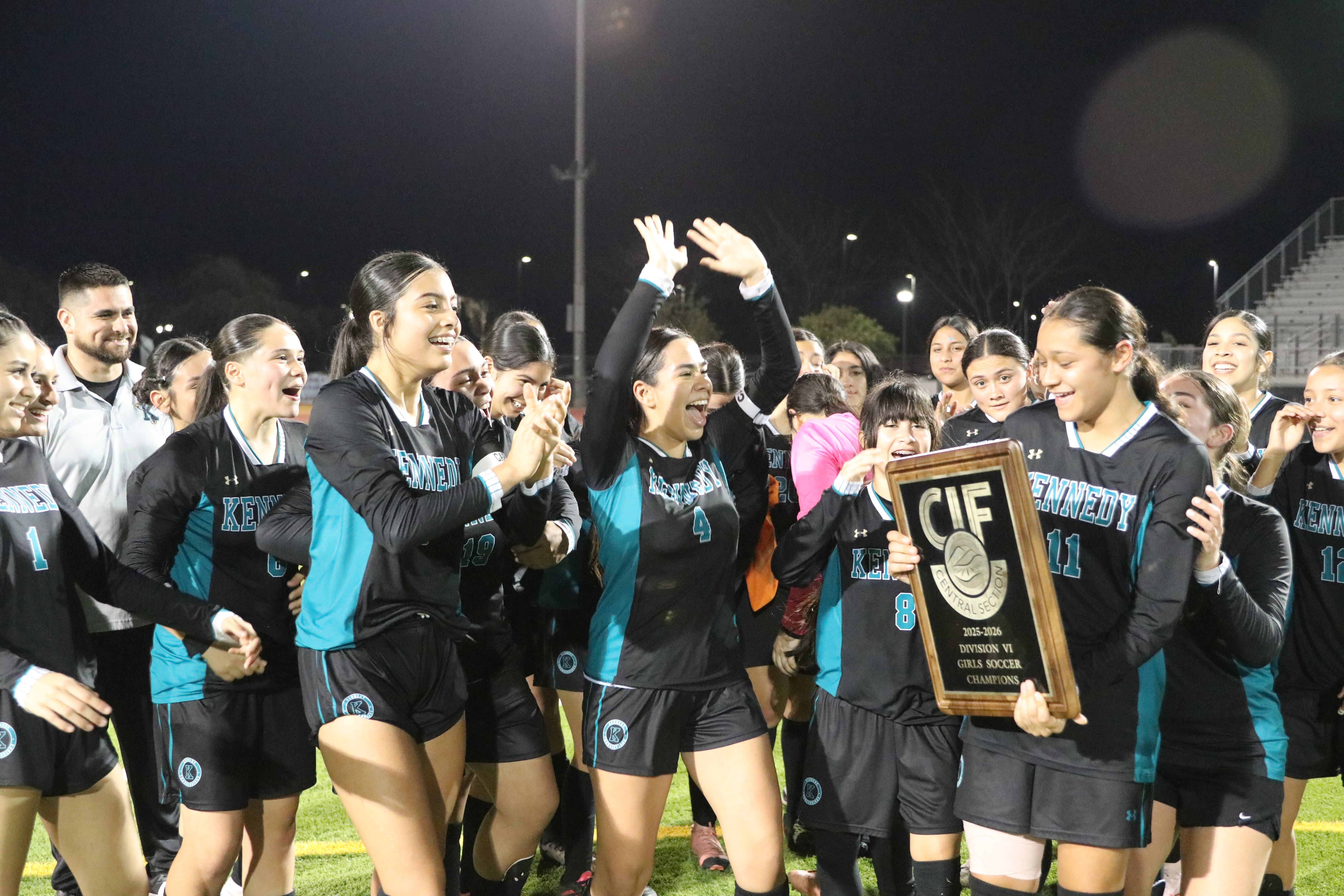 A group of people, likely a sports team, celebrating their victory while holding a trophy in the foreground, with a night-time setting and stadium lights in the background.