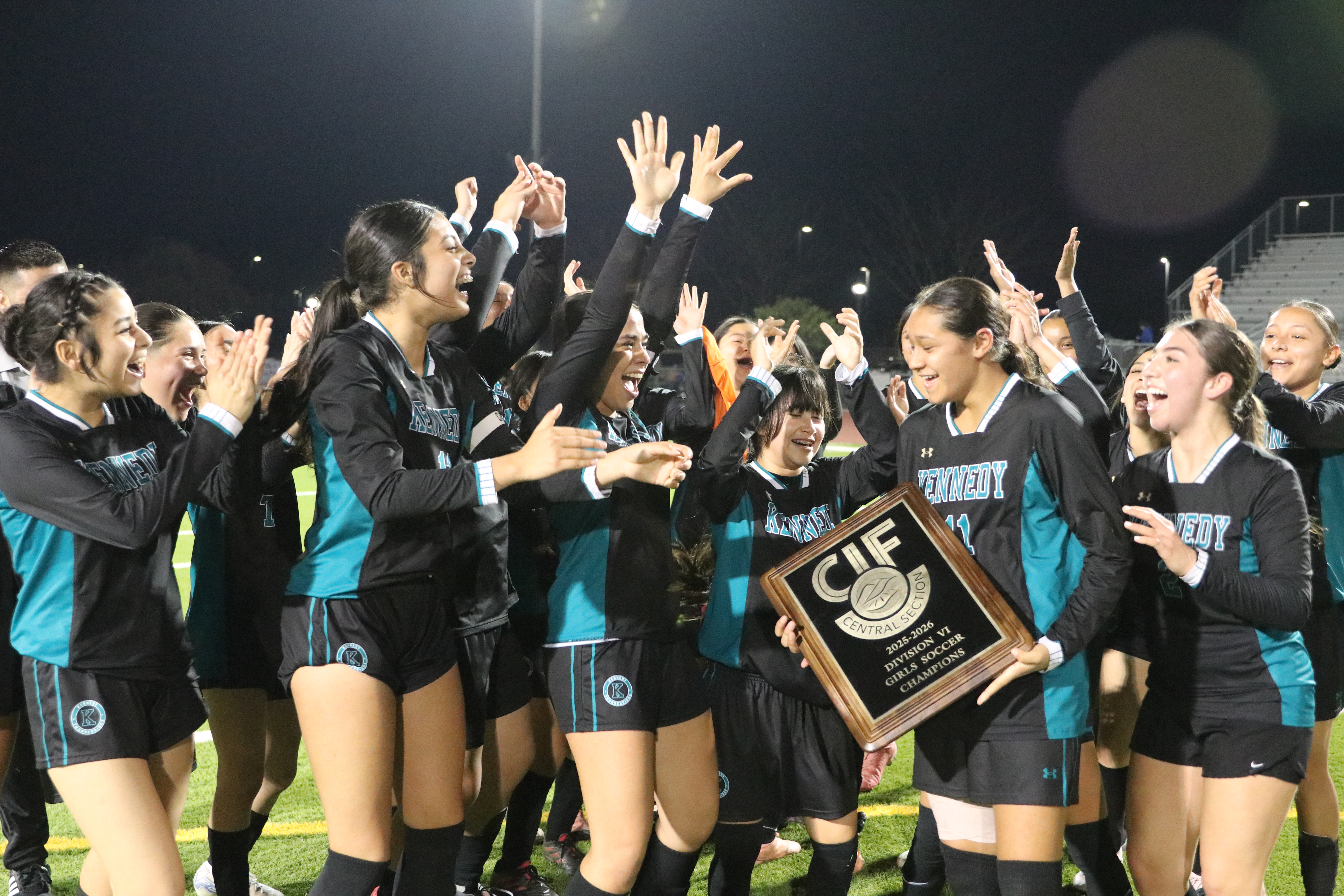 A group of young women in sports uniforms are celebrating and raising their hands in the air, holding a trophy, on a sports field at night.