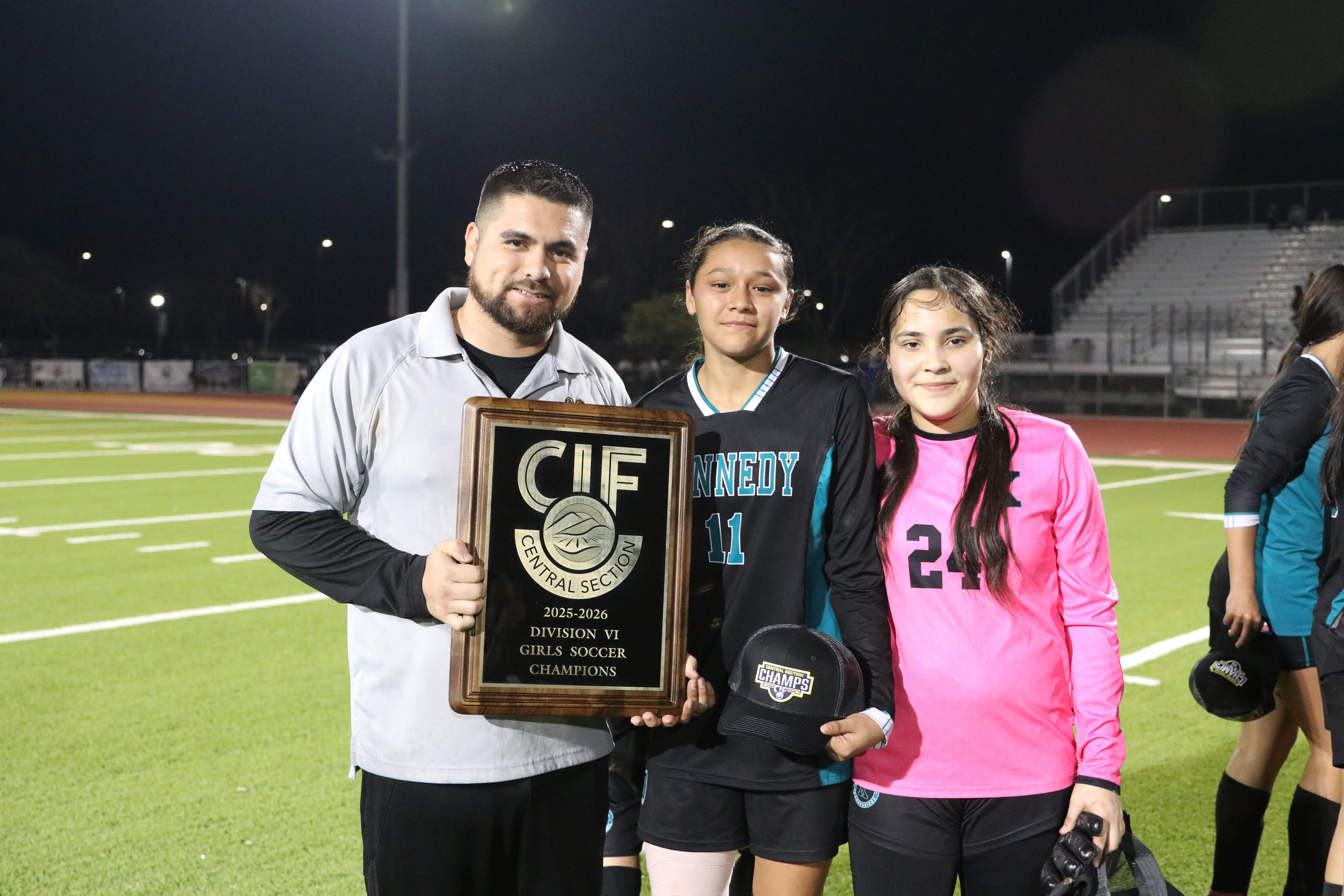 The image shows a man and two young women holding a trophy on a sports field at night, with stadium lights illuminating the scene.