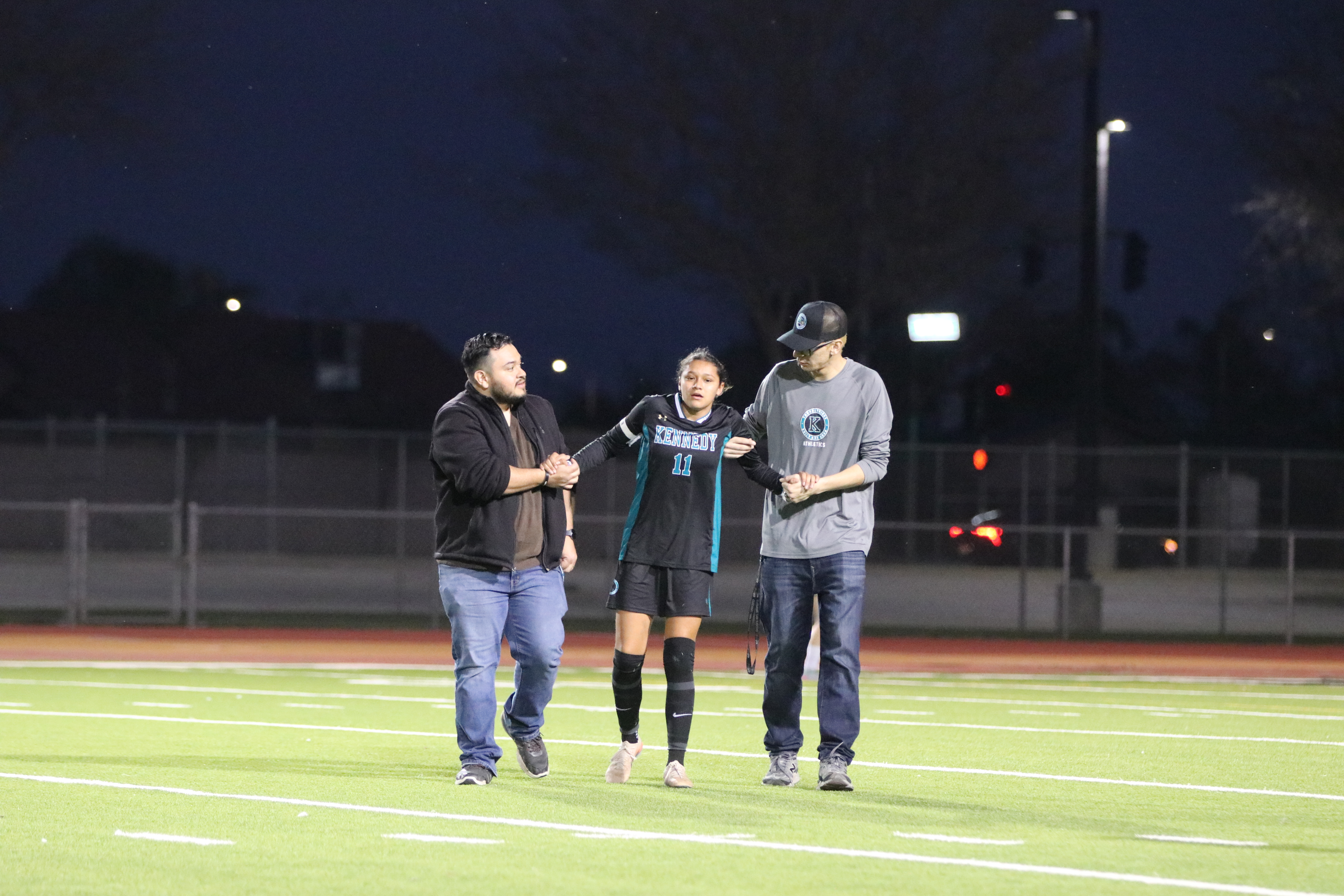 Three people, two men and one woman, are walking together on a sports field at night, with lights and a fence visible in the background.