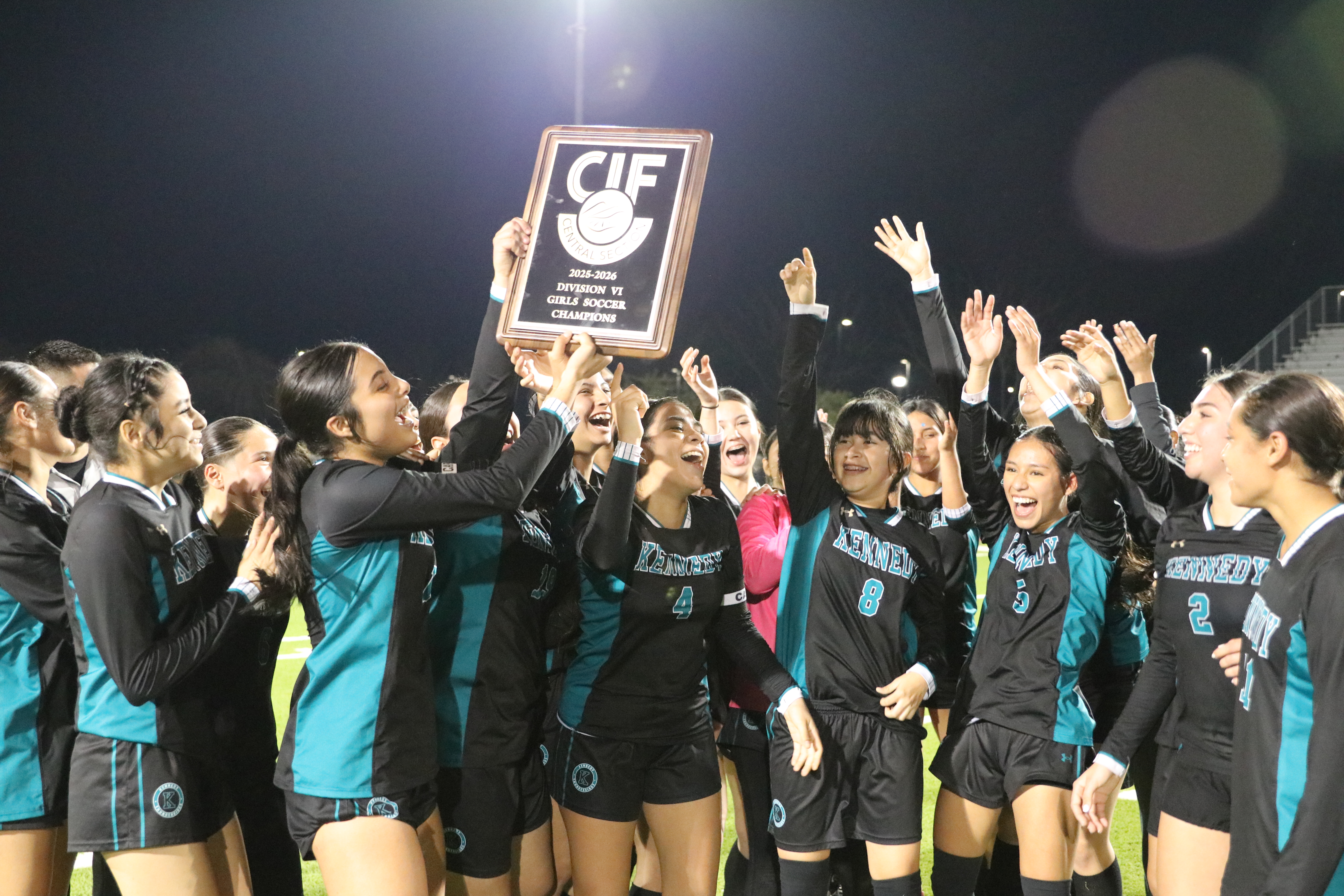 A group of people, likely a sports team, are gathered together and celebrating, holding up a sign or trophy in the foreground, with a dark background.