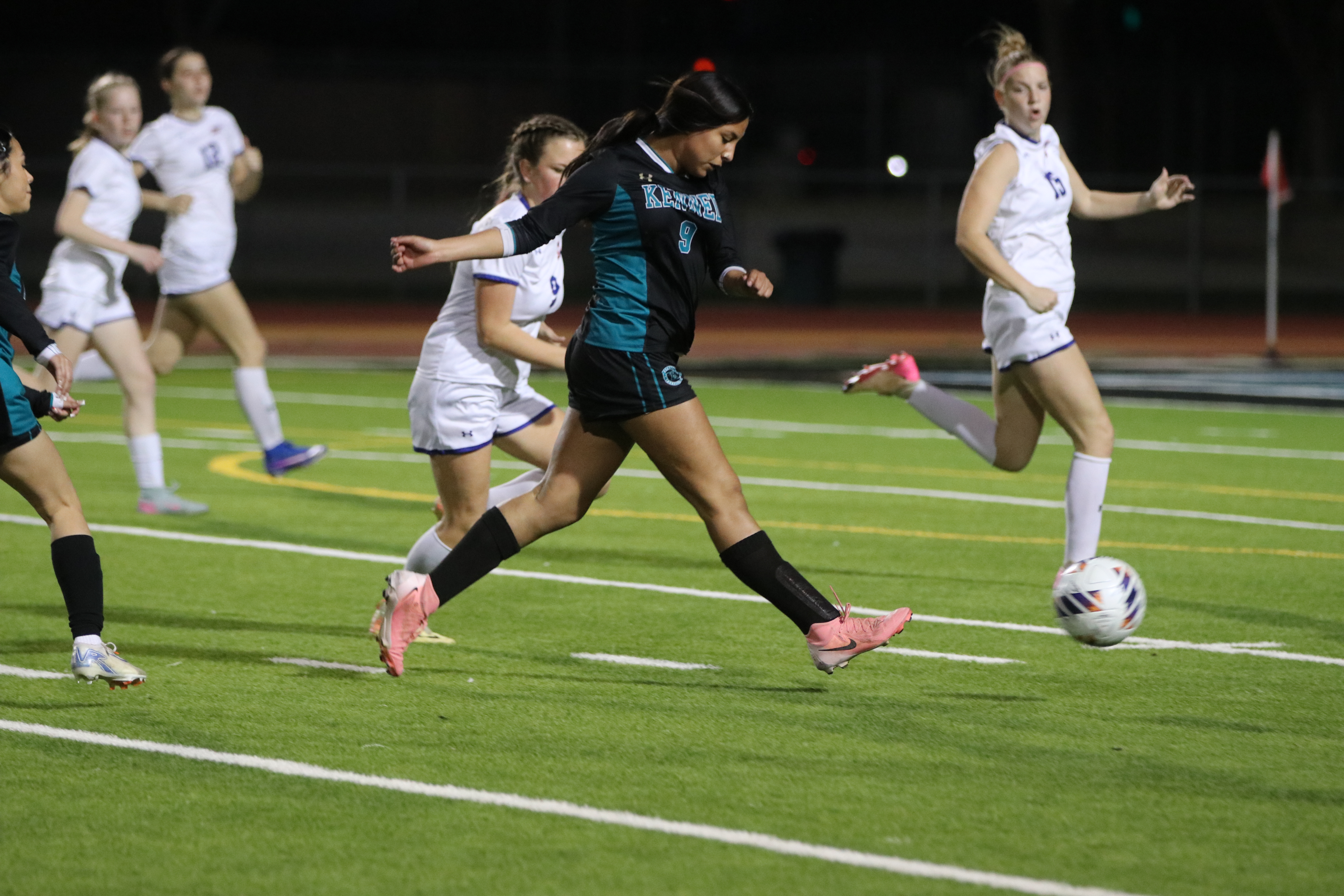 The image shows a group of female soccer players on a field, with one player in the foreground dribbling the ball down the field while her teammates surround her.