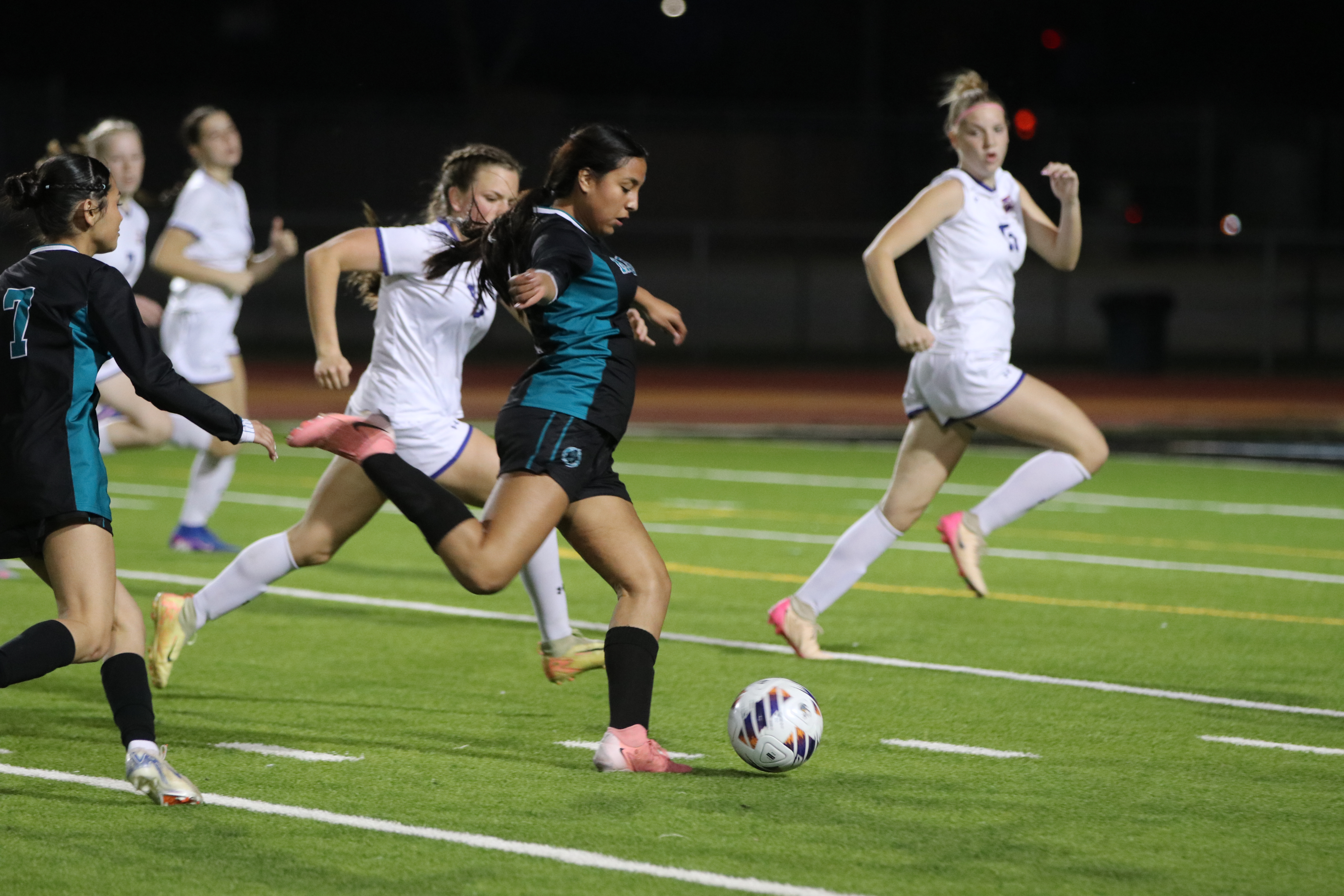 The image shows a group of female soccer players competing on a well-lit soccer field at night, with the players in the foreground engaged in an intense moment of the game.