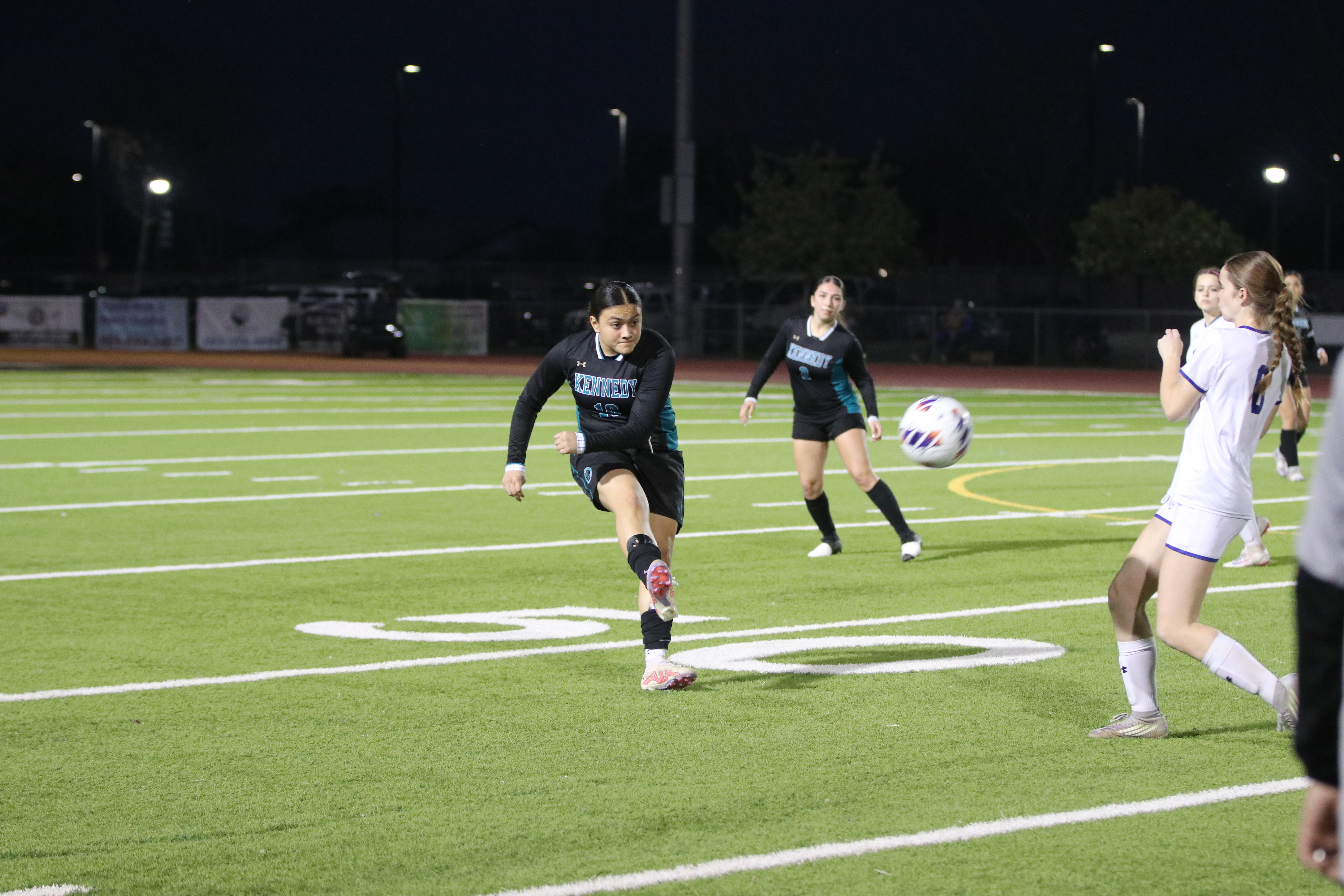The image shows a group of people playing soccer on a well-lit soccer field at night, with the field's markings and goal posts visible in the background.