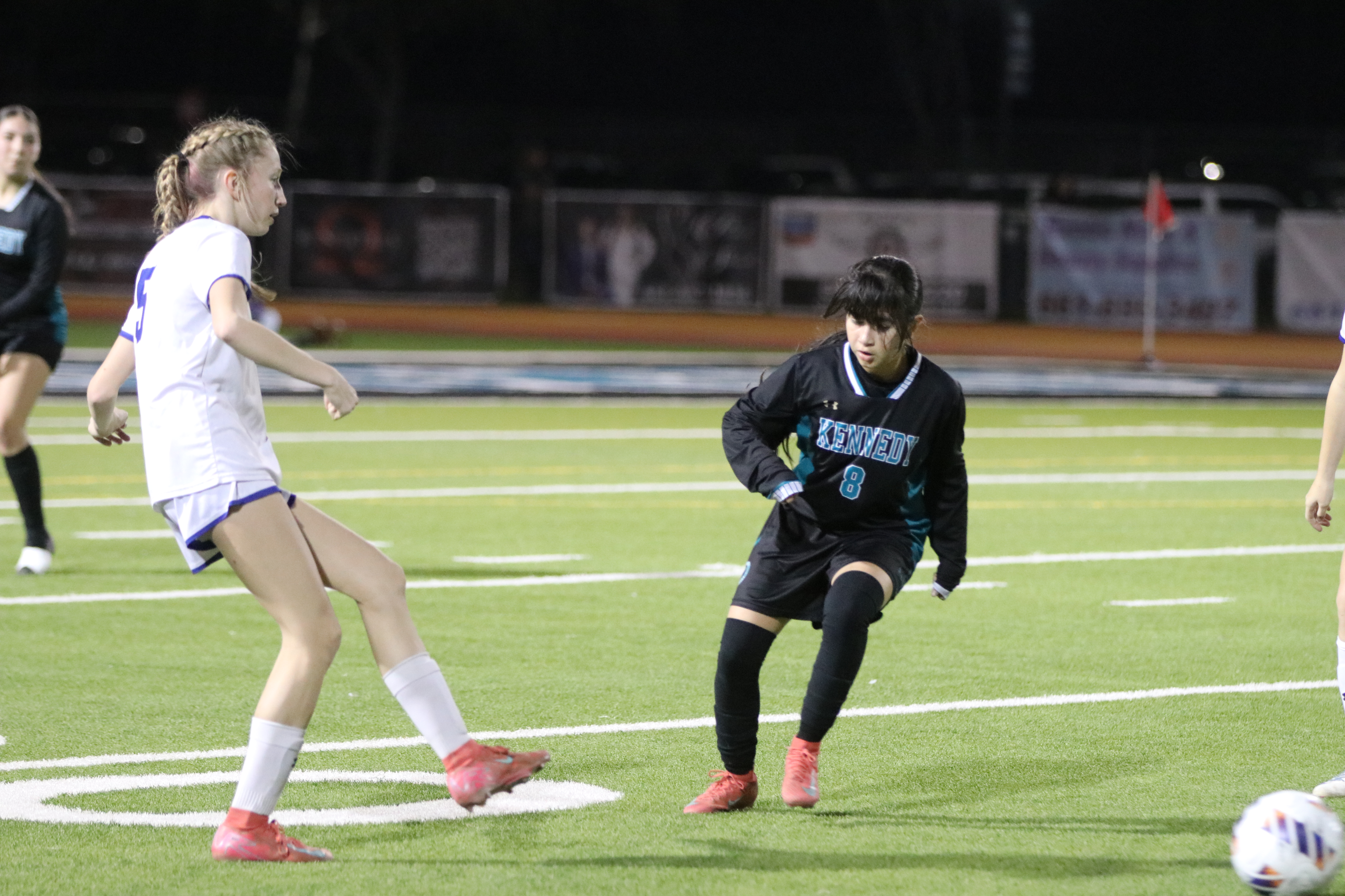 Two female soccer players are engaged in a match on a well-lit soccer field, with spectator stands visible in the background.