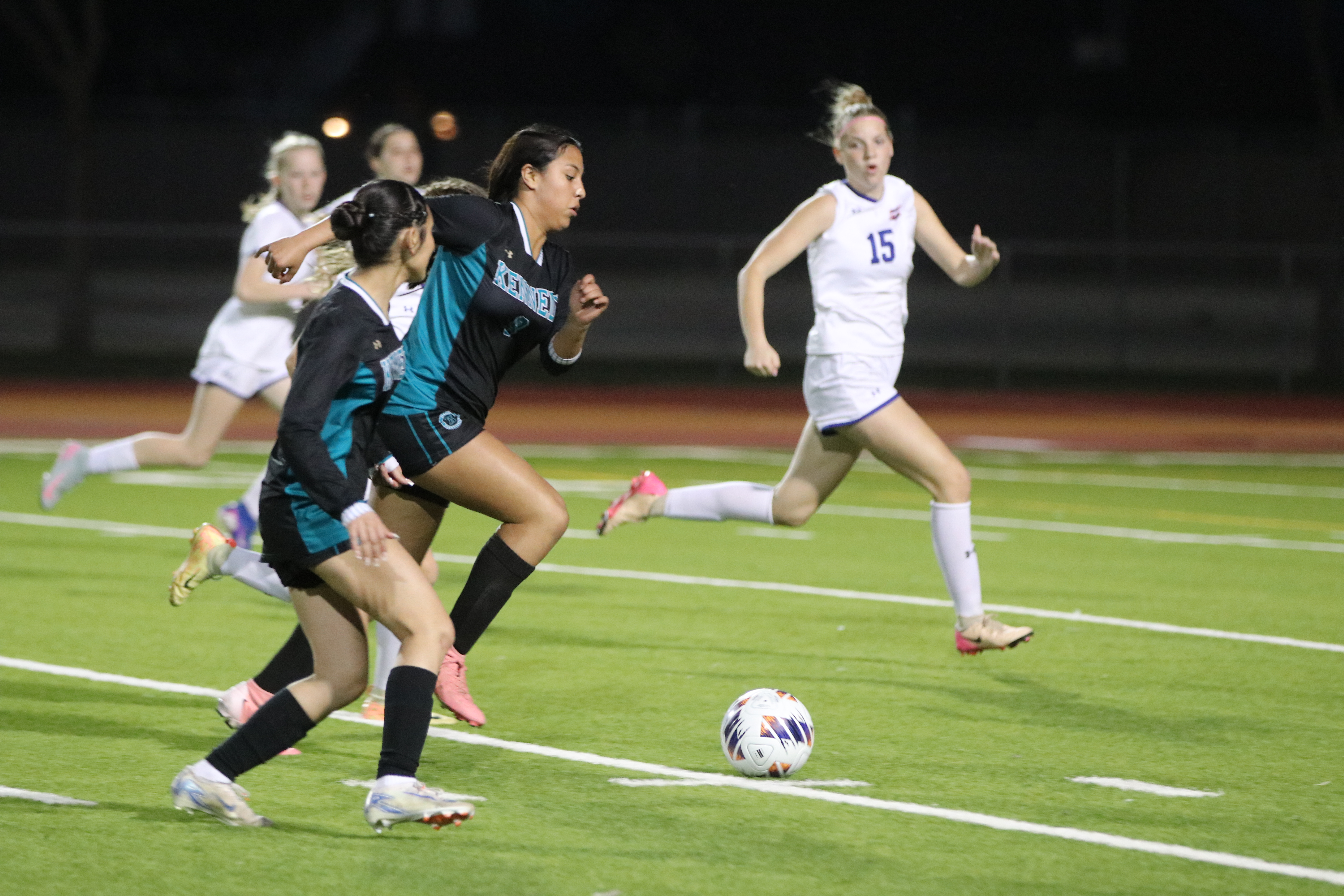 The image depicts a soccer game taking place on a lighted field at night, with players in white and blue uniforms competing for the ball.
