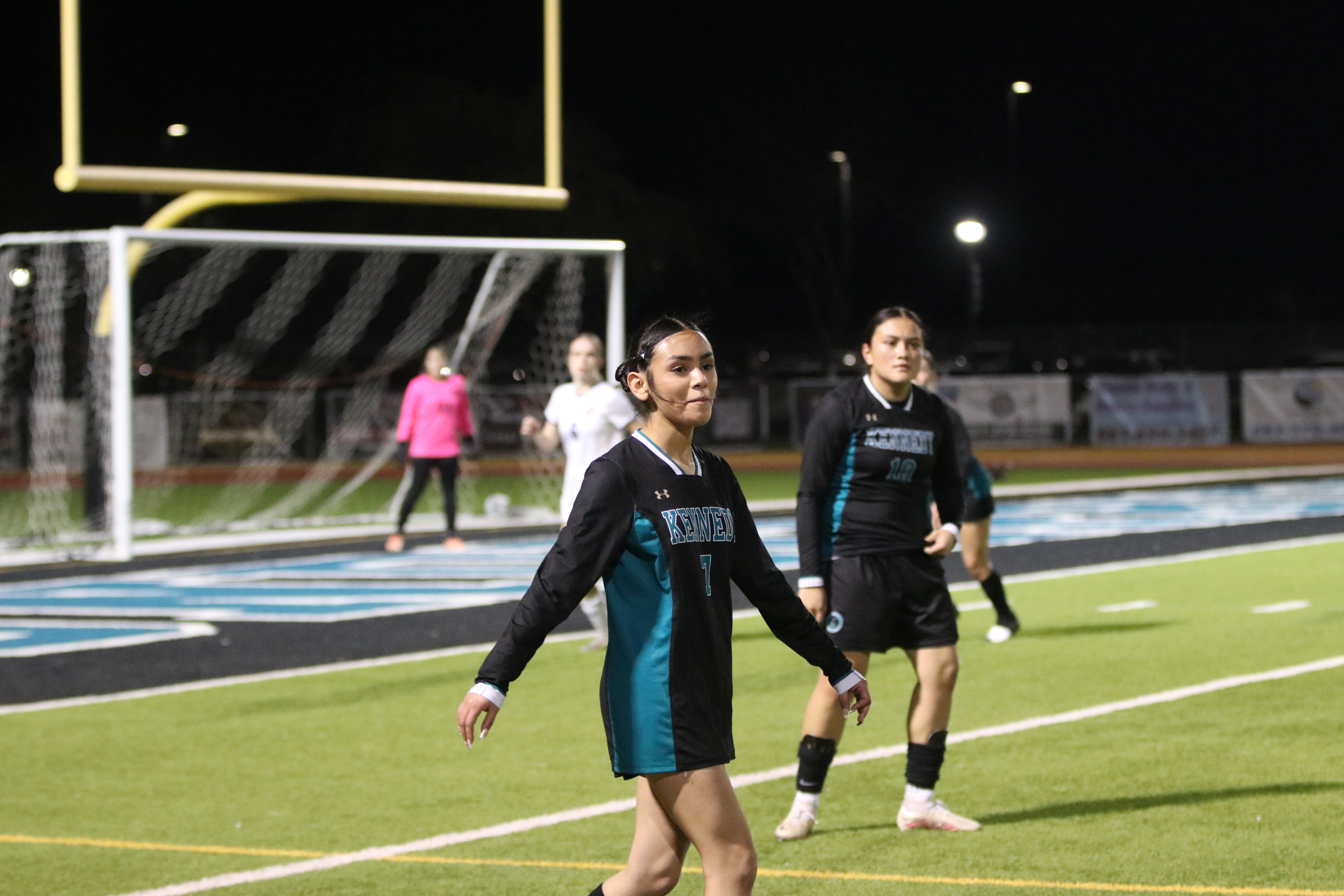 The image shows two people, a woman and a man, standing on a sports field at night, with a goal post and lights visible in the background.