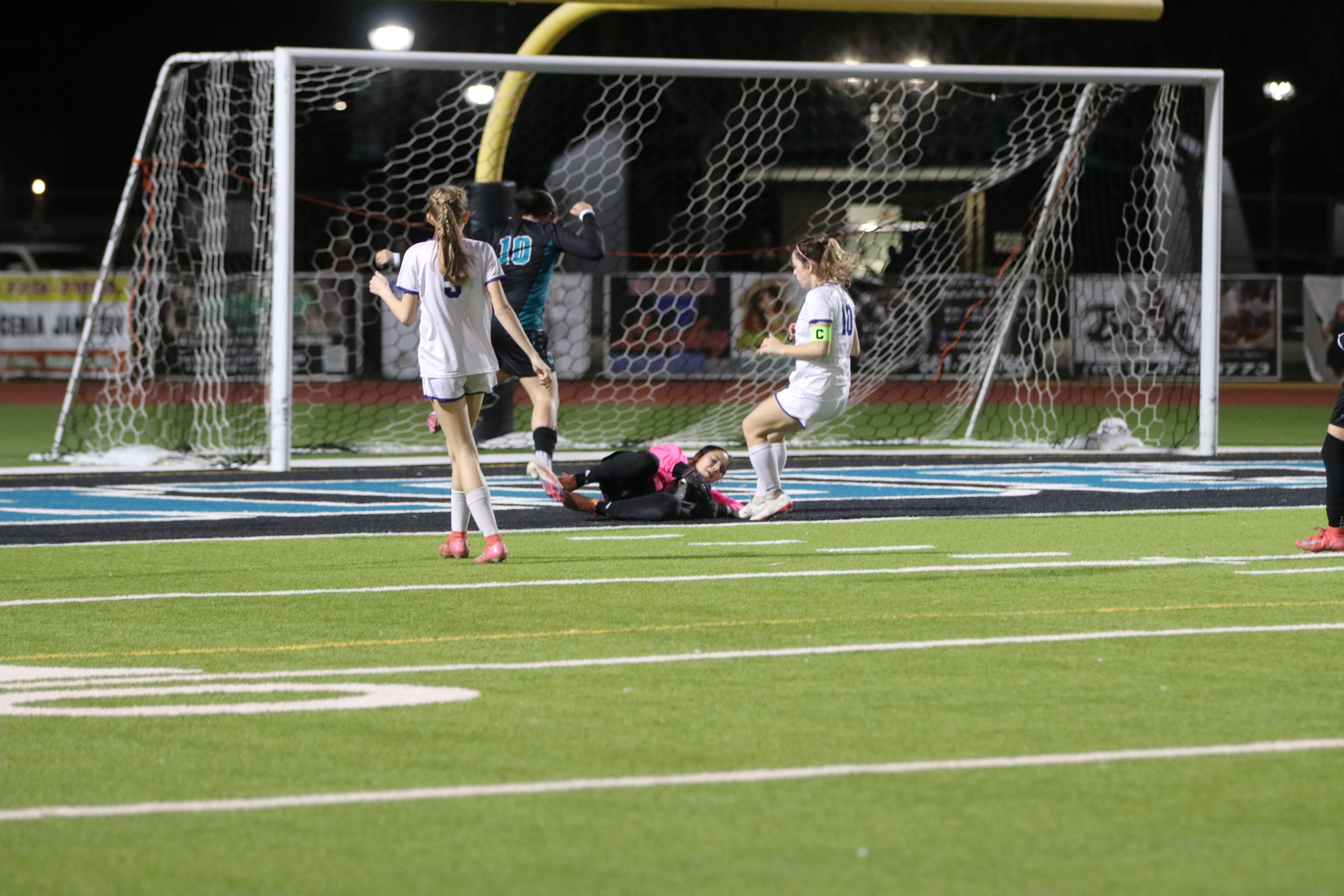 The image shows a soccer game taking place on a well-lit field, with players in white and blue uniforms competing near the goal posts.