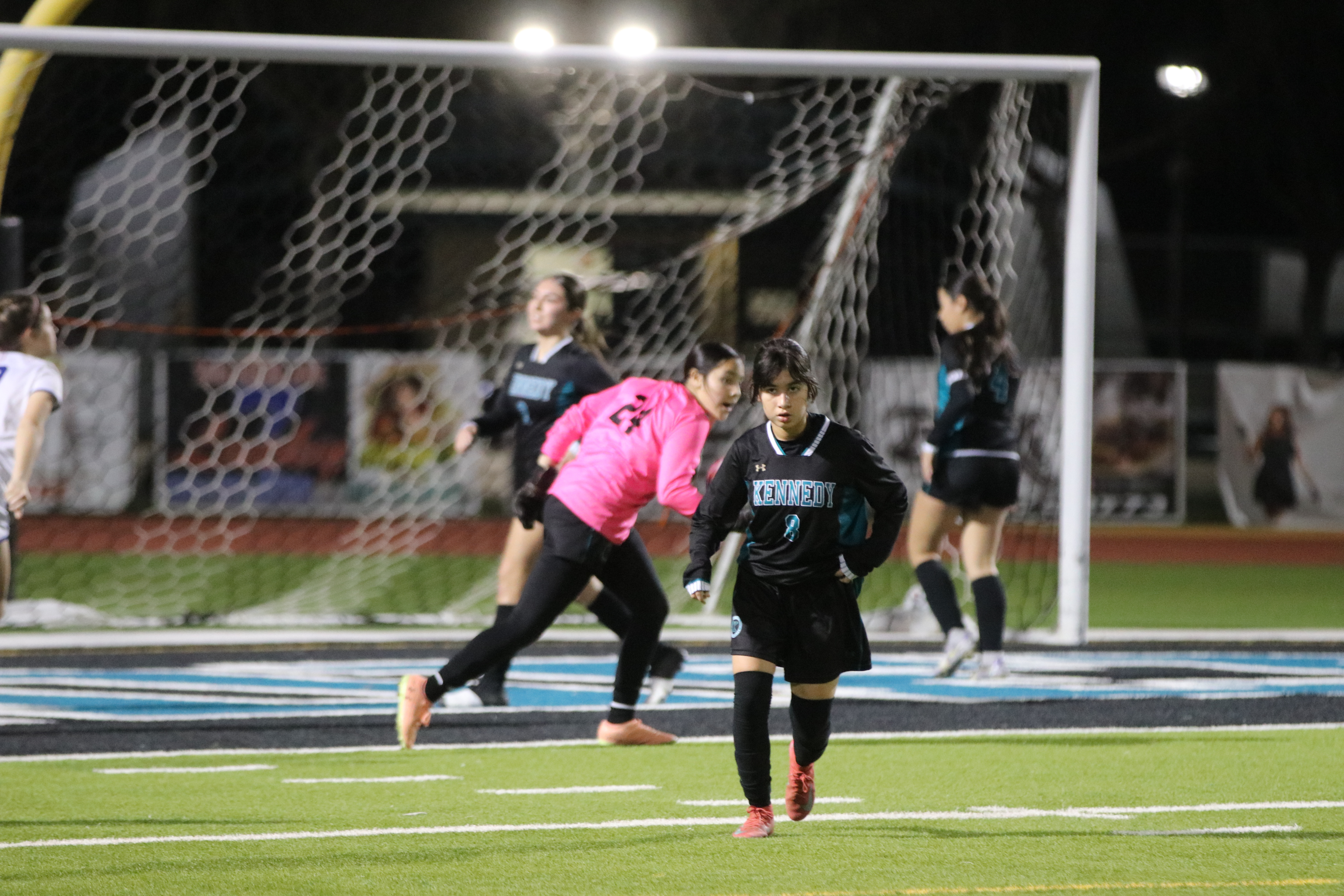 The image shows a group of soccer players on a field, with a goalkeeper in the foreground and other players in the background near the goal.