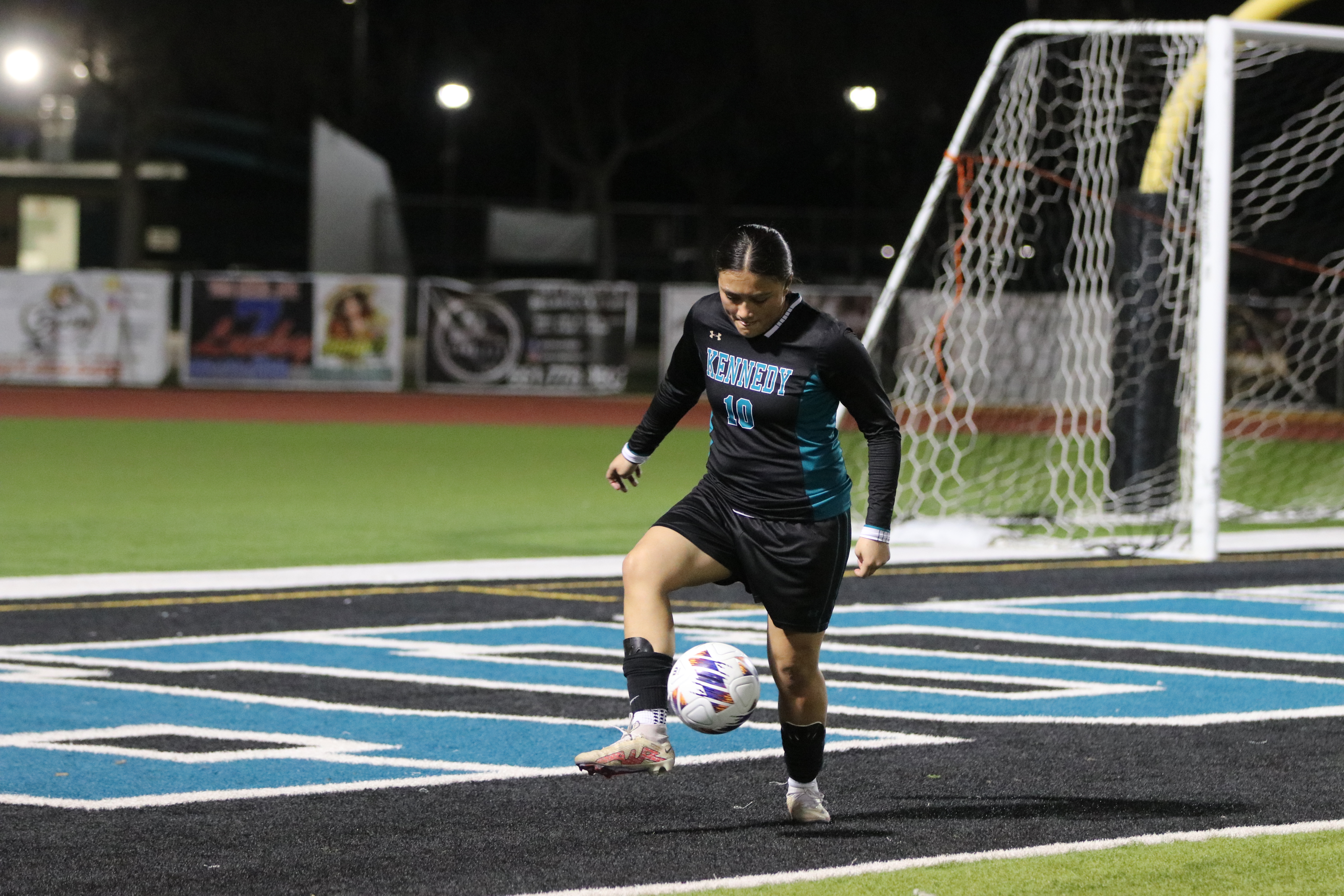 A soccer player in a black uniform is dribbling the ball on a brightly lit artificial turf field, with advertising boards and lights visible in the background.