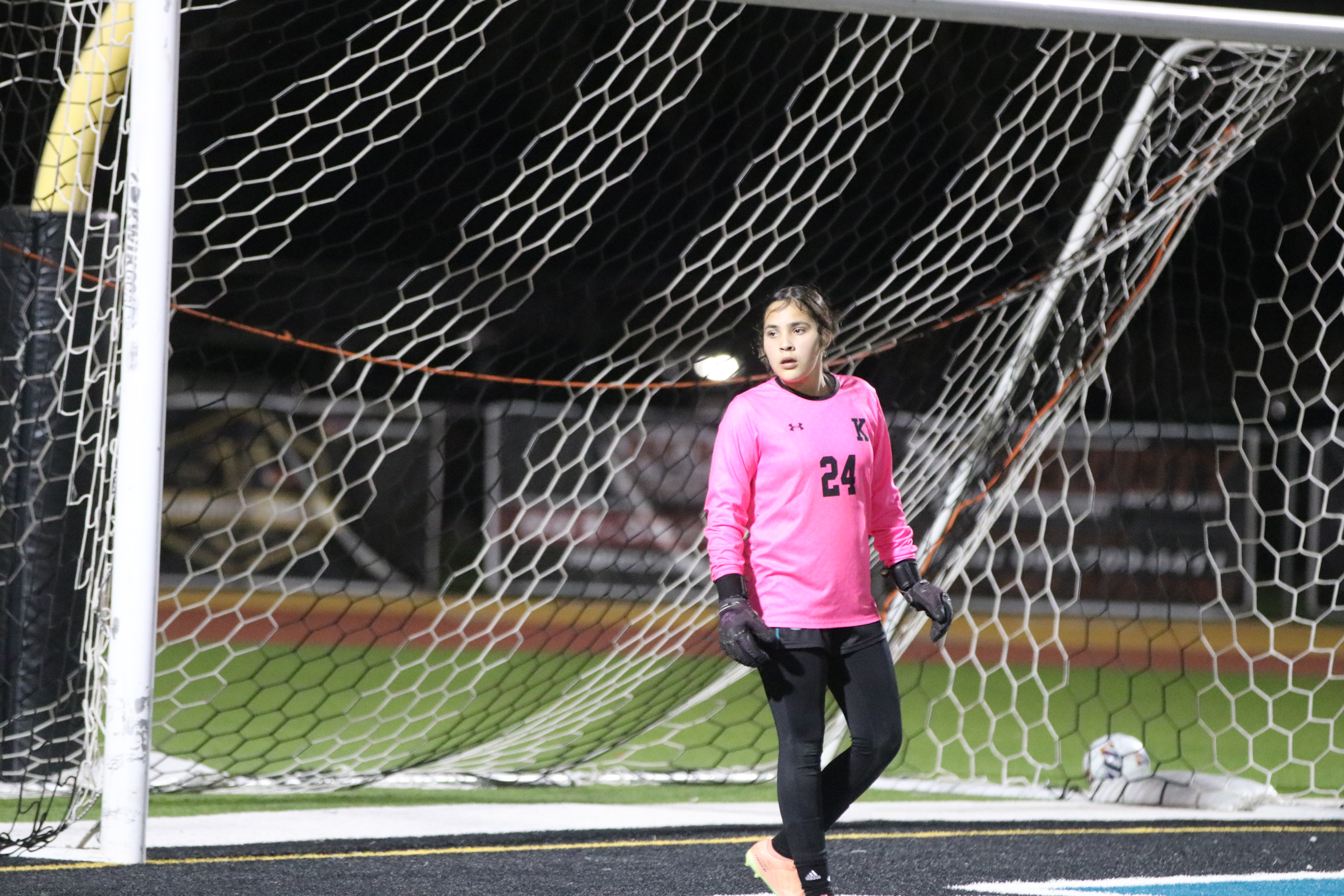 A young female soccer player in a pink jersey stands in a soccer goal, with a black net and field visible in the background.