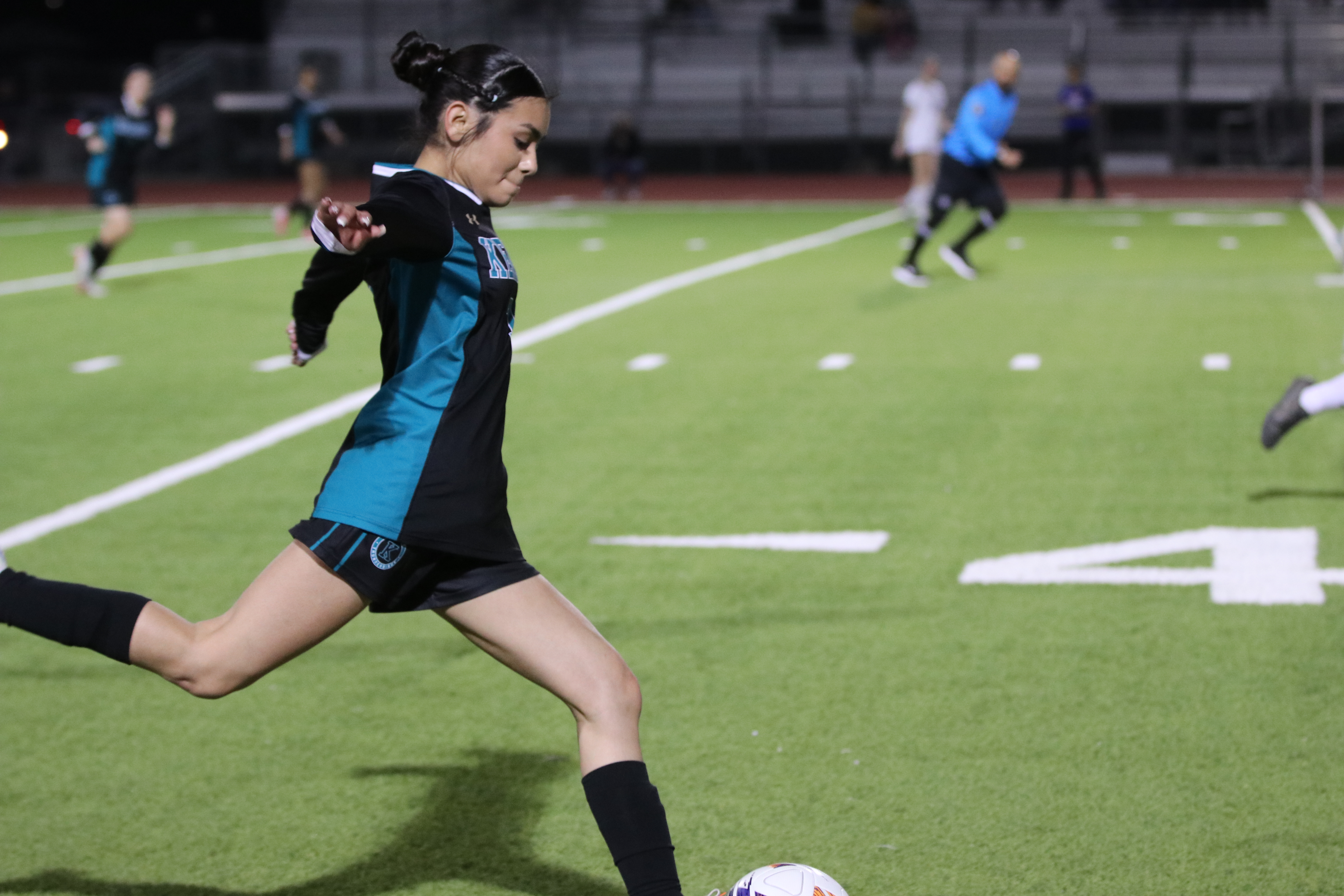 A young woman in a blue and black uniform is running with a soccer ball on a well-lit sports field, with other players visible in the background.