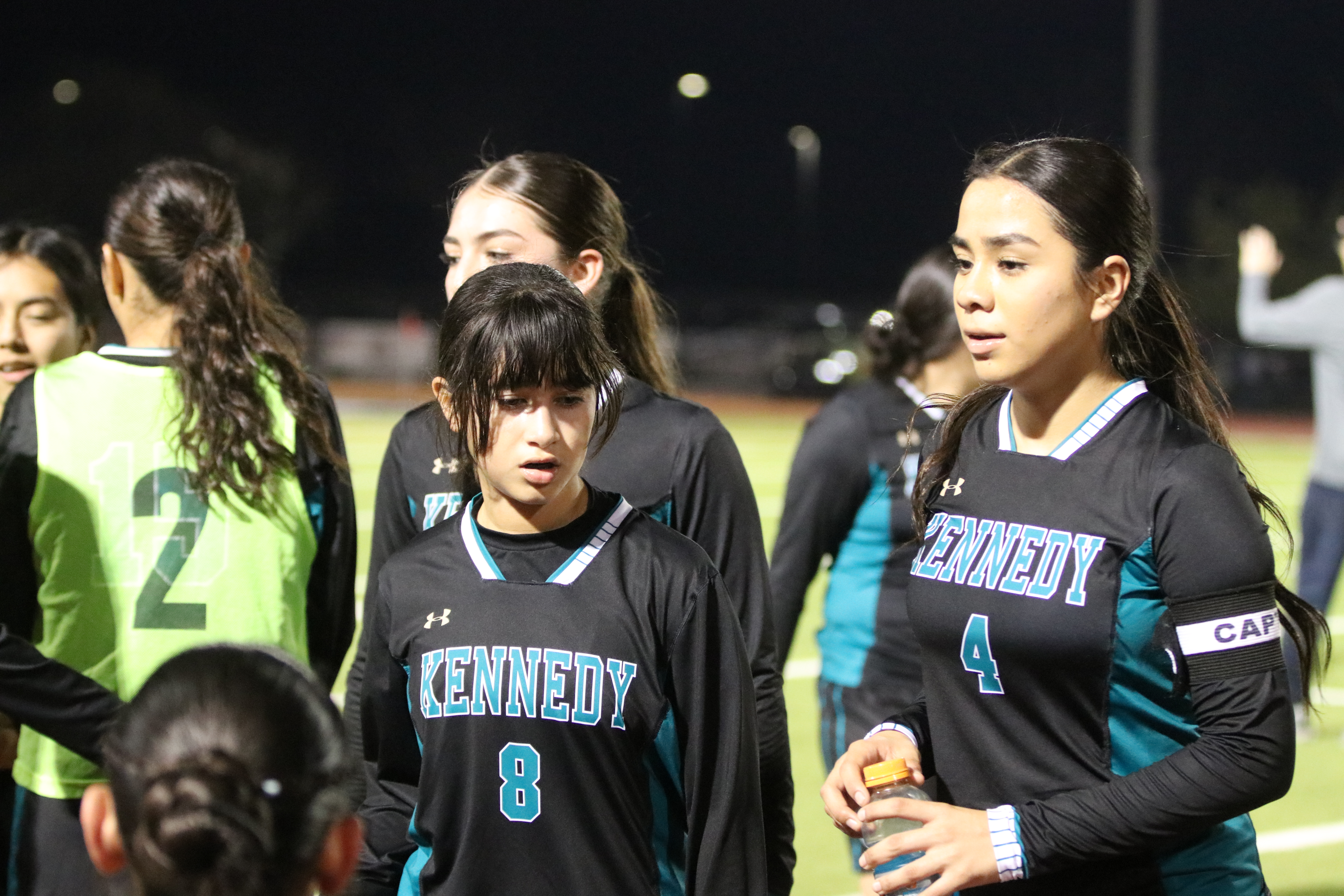The image shows a group of young female athletes in sports uniforms, standing together on a sports field at night with lights in the background.