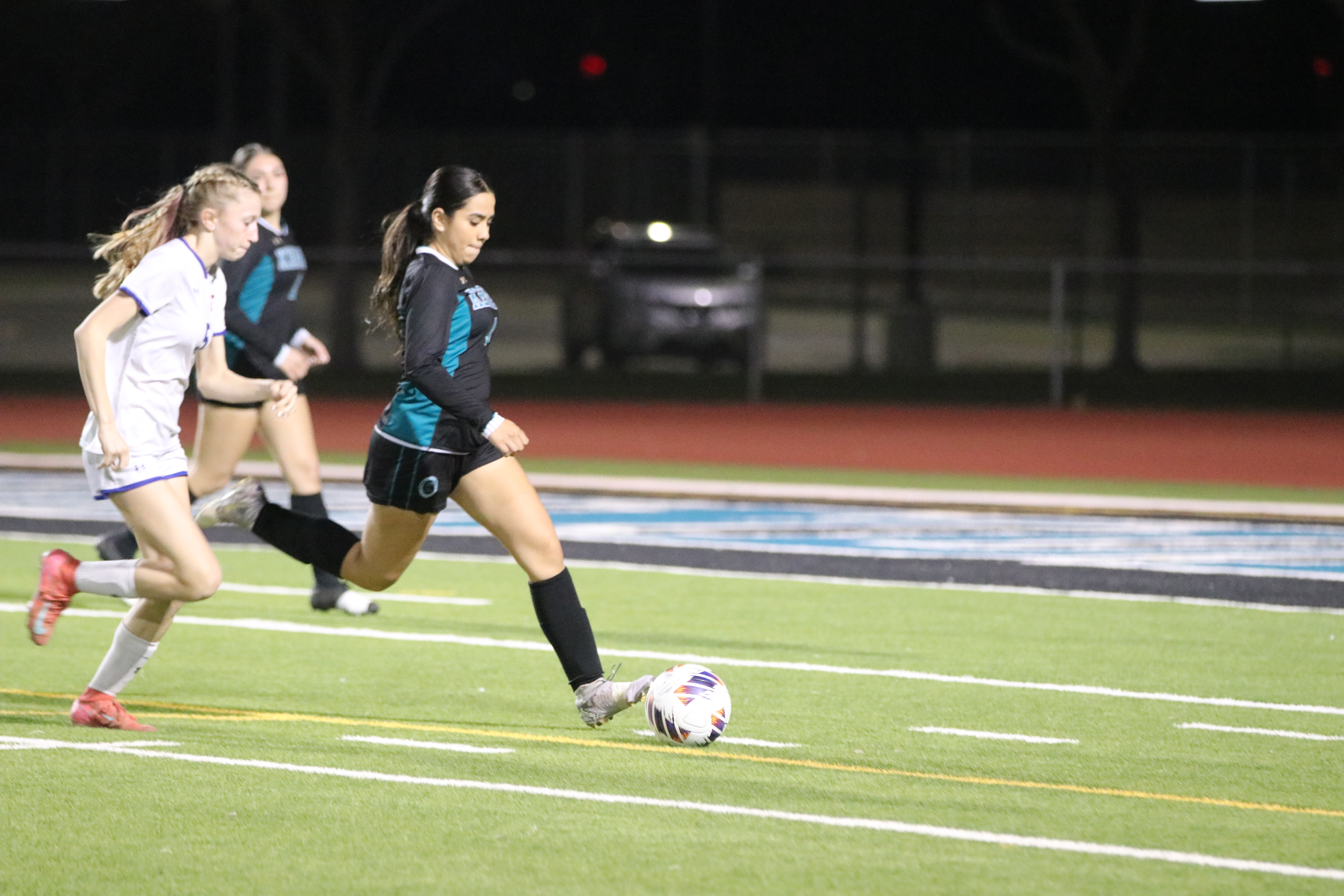 Two female soccer players are running on a soccer field, with a soccer ball between them. In the background, a car can be seen parked near the field.