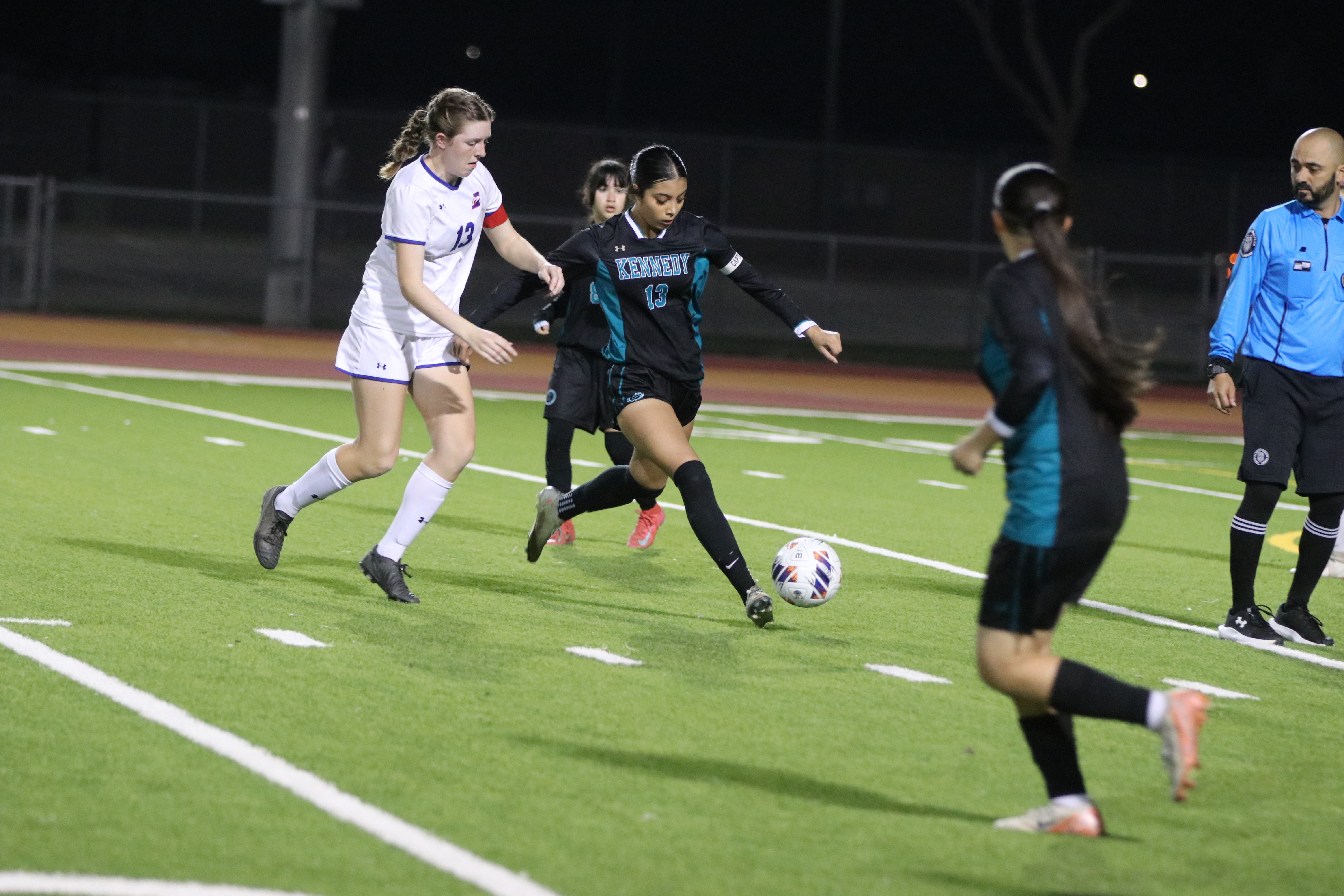 The image depicts a soccer game taking place on a lighted field at night, with players from both teams actively engaged in the match.