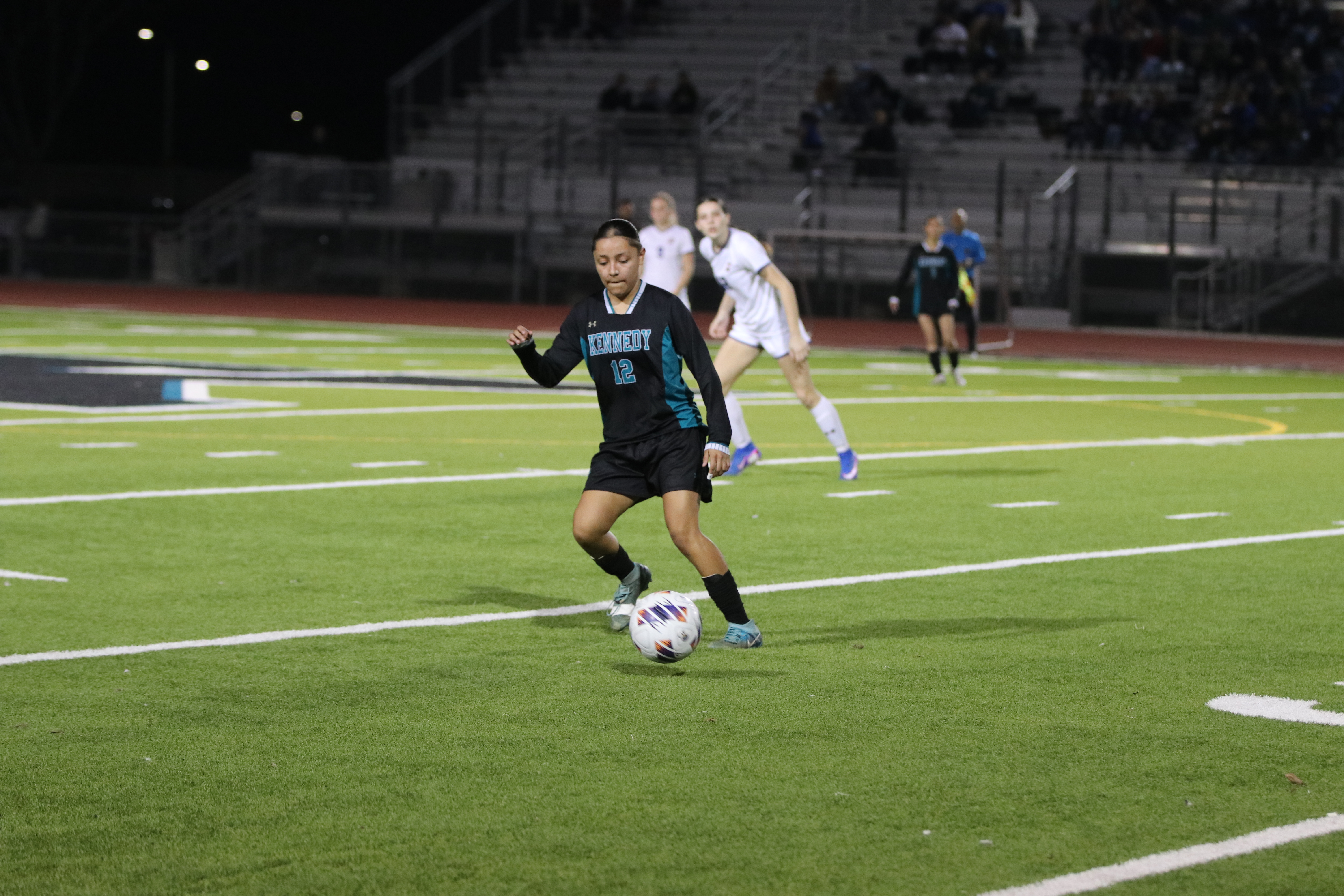 A soccer player in a black jersey is dribbling the ball down the field, with other players visible in the background on a lighted soccer field.