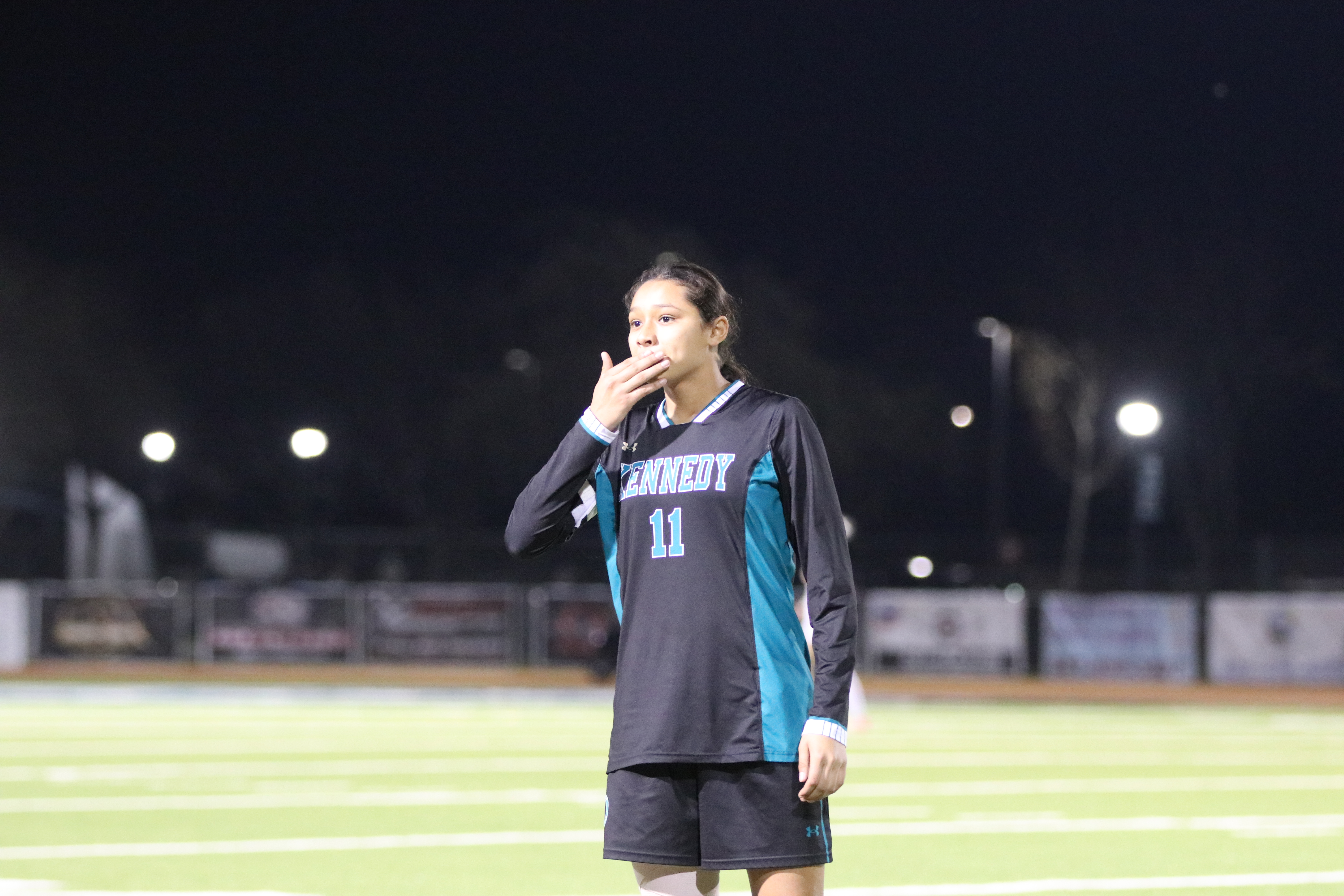 A person wearing a gray and teal jersey stands on a brightly lit sports field at night, with lights and bleachers visible in the background.