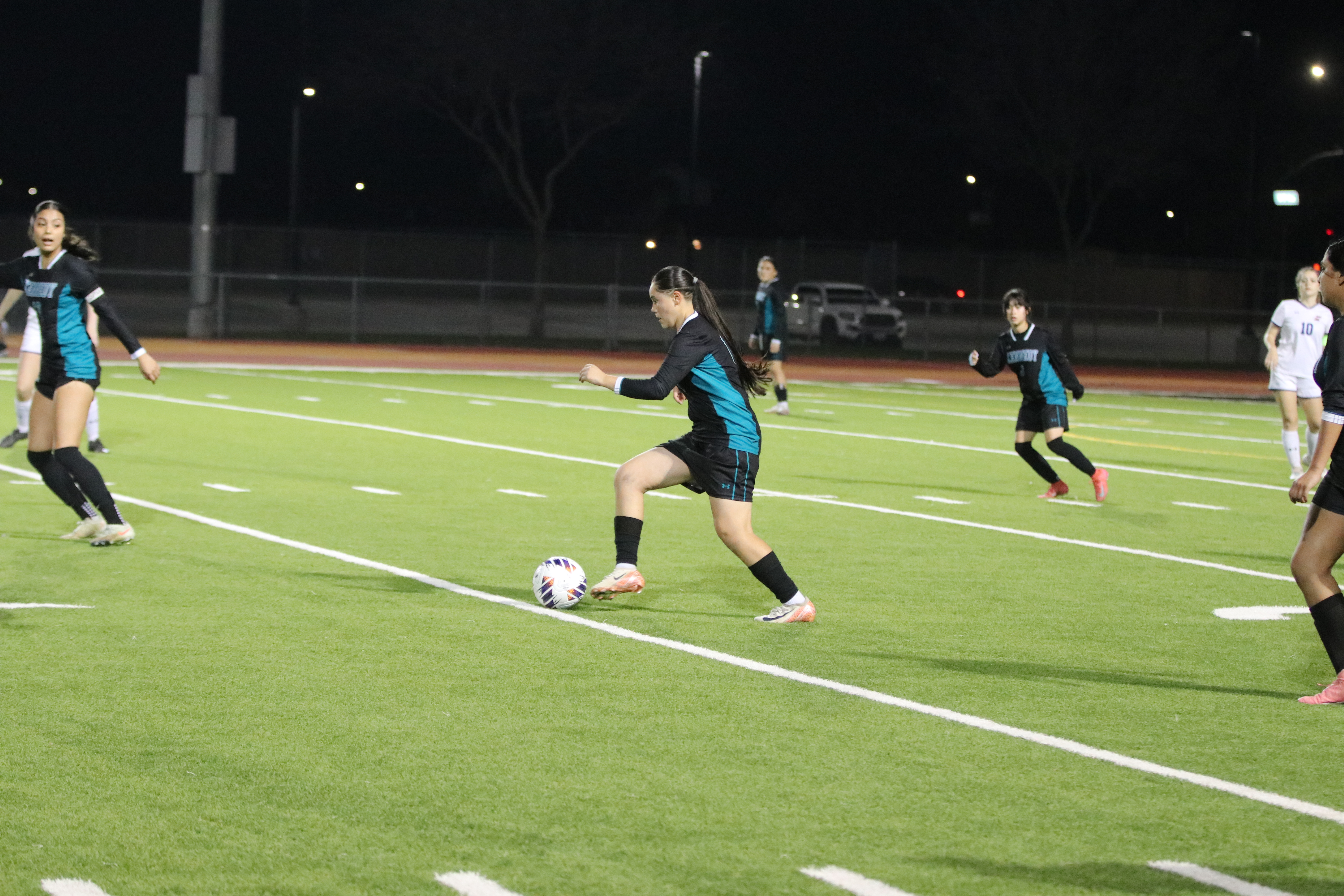 The image shows a soccer game being played on a well-lit field at night, with players in blue and white uniforms engaged in the action.