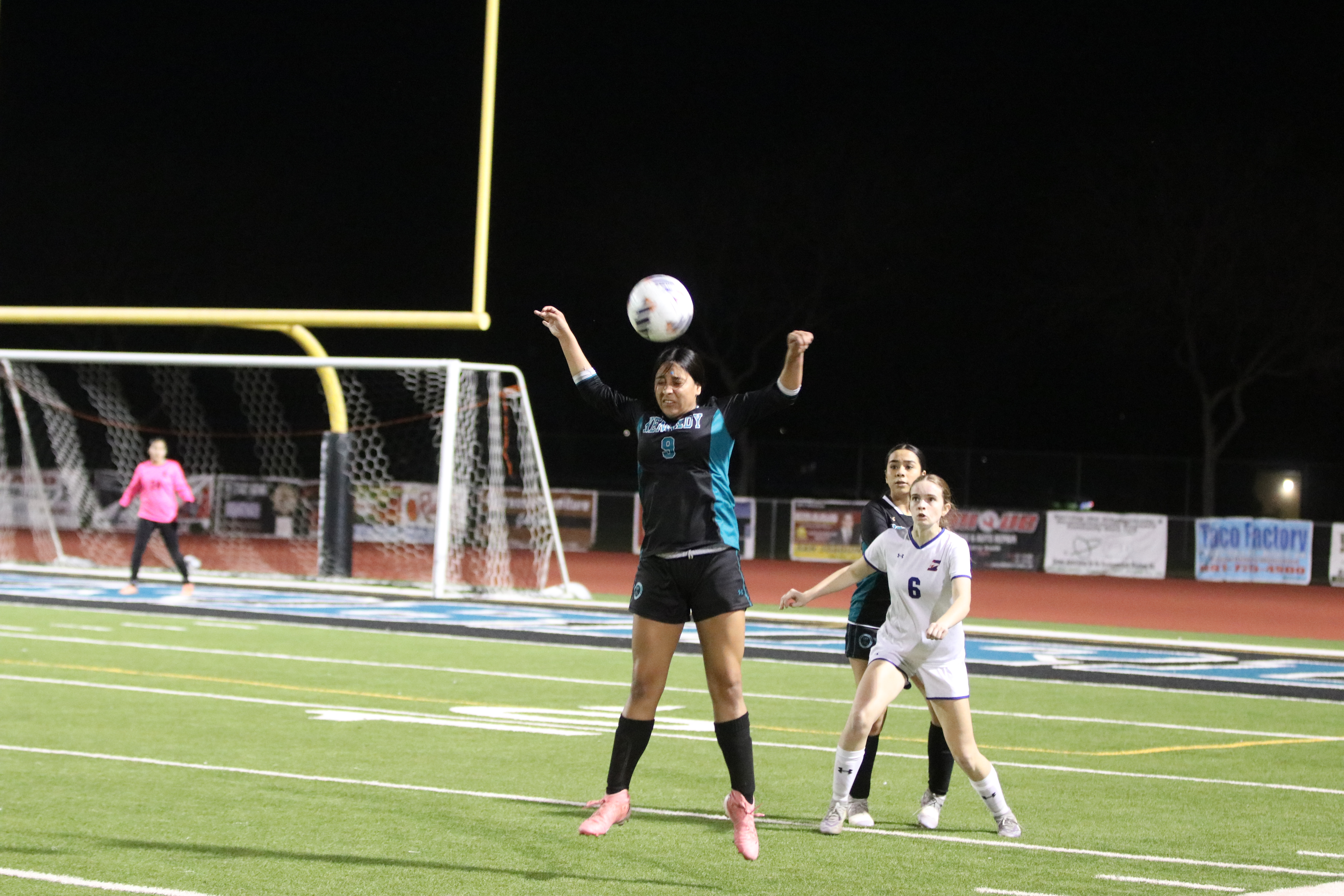 The image shows two female soccer players competing on a brightly lit soccer field at night, with one player reaching up to head the ball while the other player attempts to block the shot.