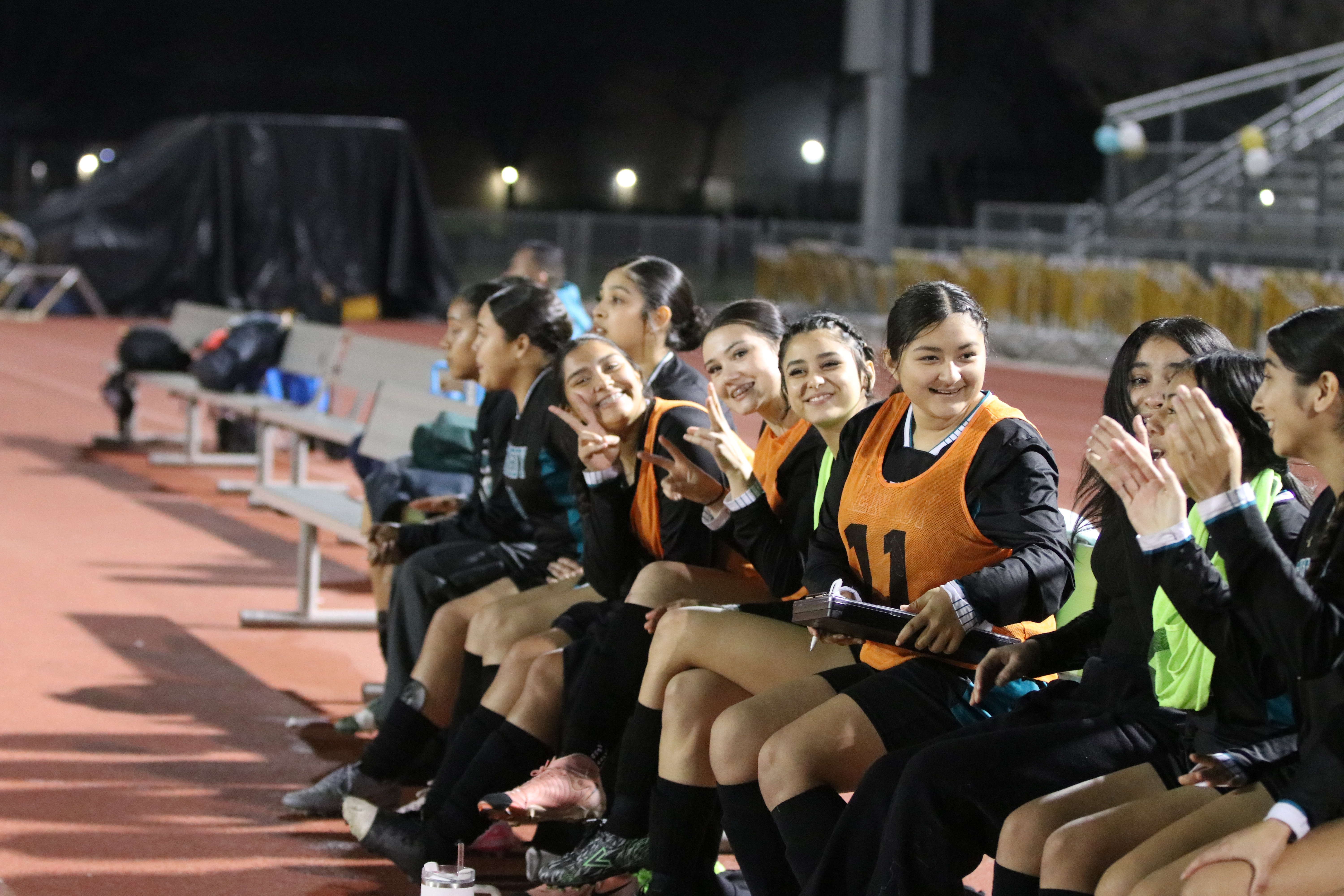A group of young women in sports uniforms sitting on a bench, with a sports field and stadium lights visible in the background.