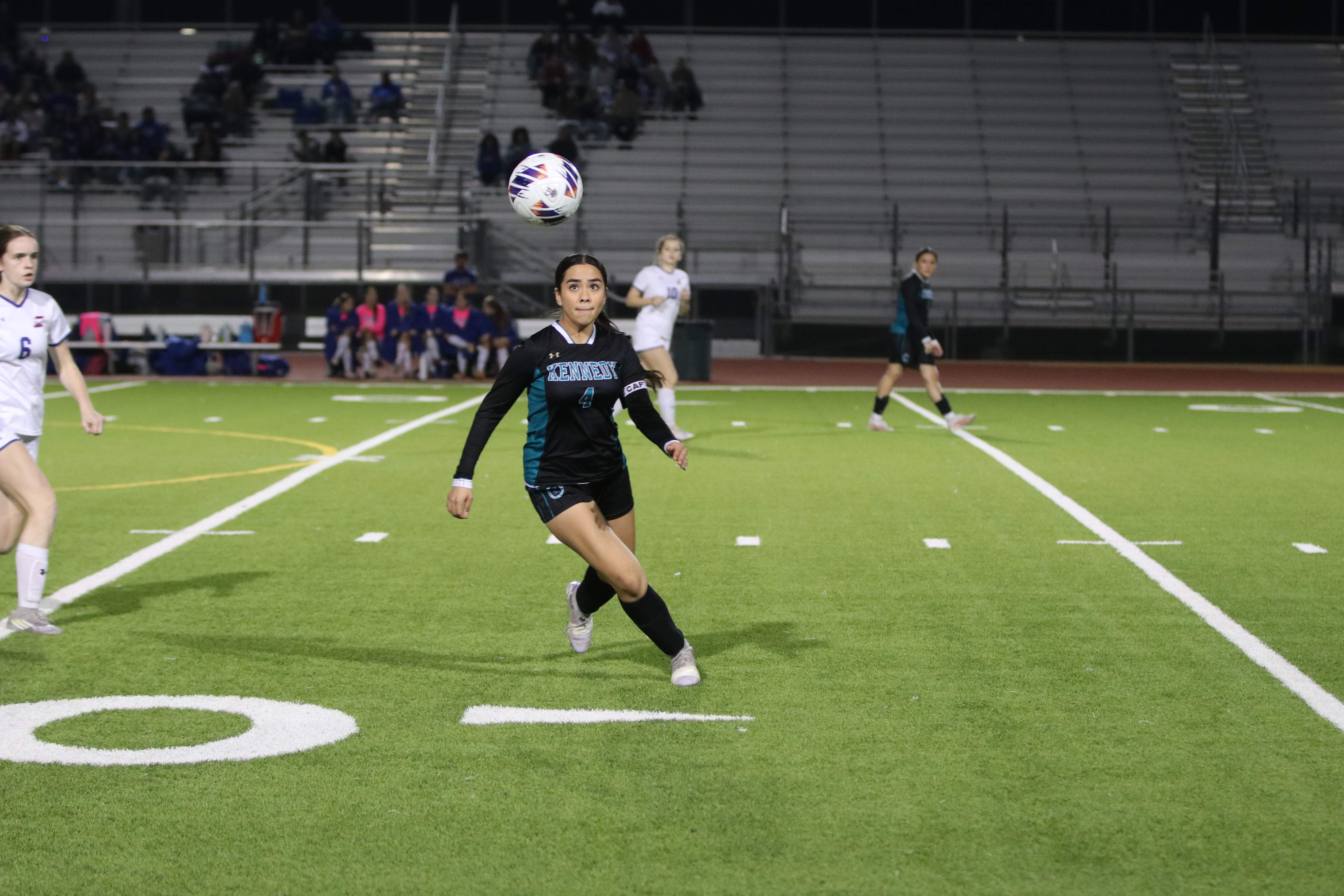 A soccer player in a black uniform is running on a brightly lit soccer field, with spectators visible in the background.