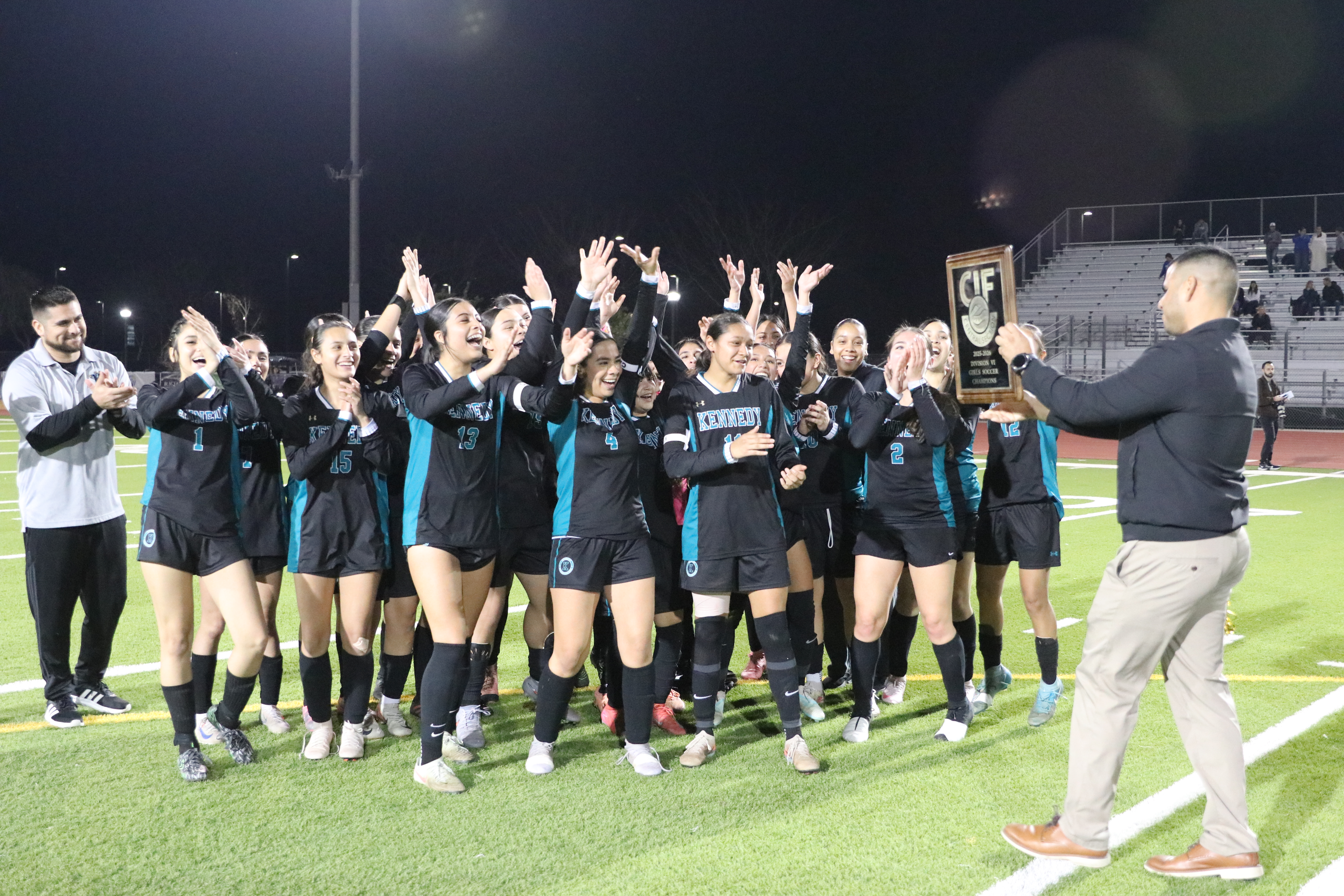 A group of soccer players, both male and female, are gathered on a soccer field at night, celebrating their victory with raised hands and cheers.