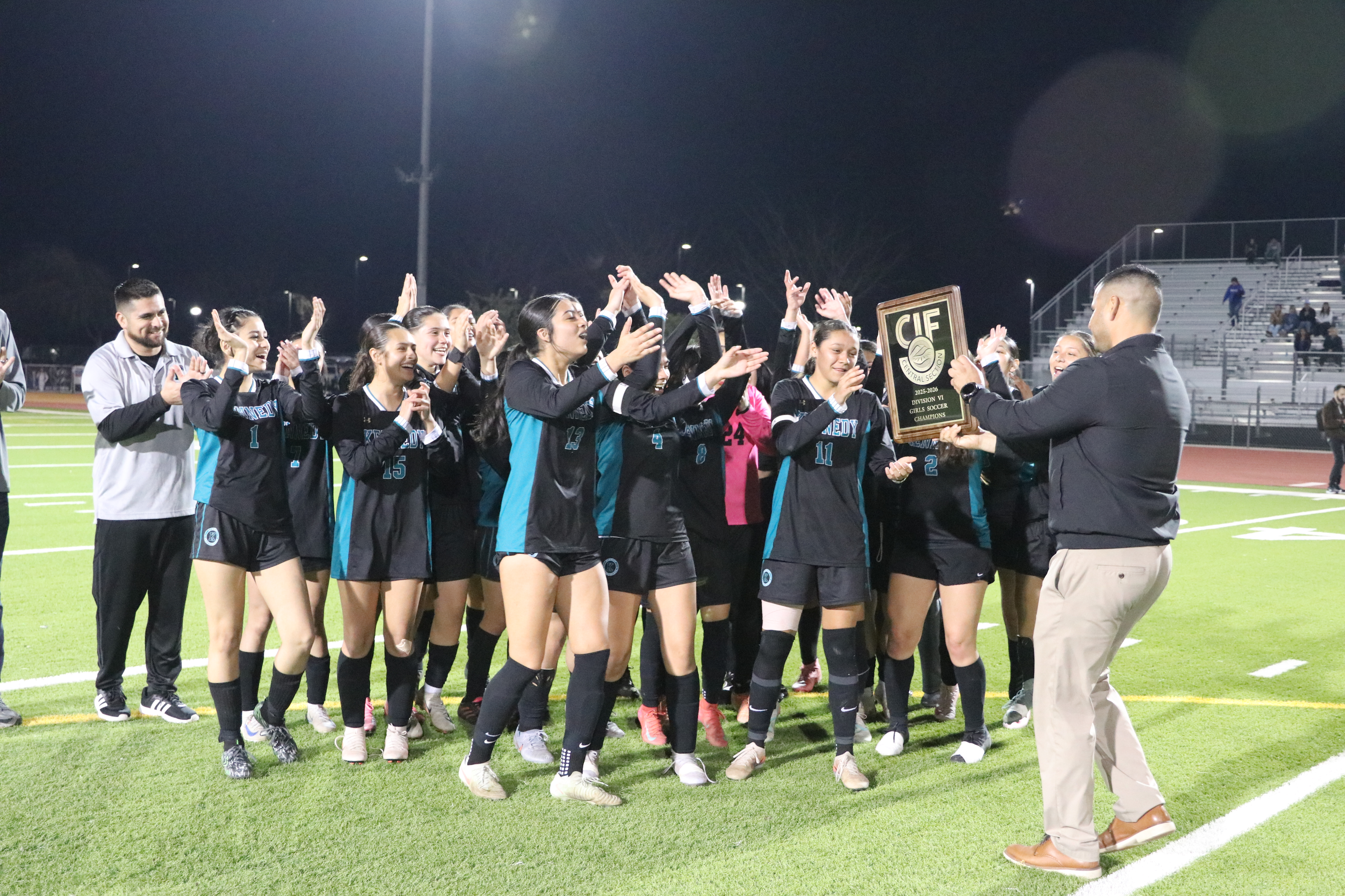 A group of people, likely a sports team, gathered together on a sports field, celebrating and holding a trophy.