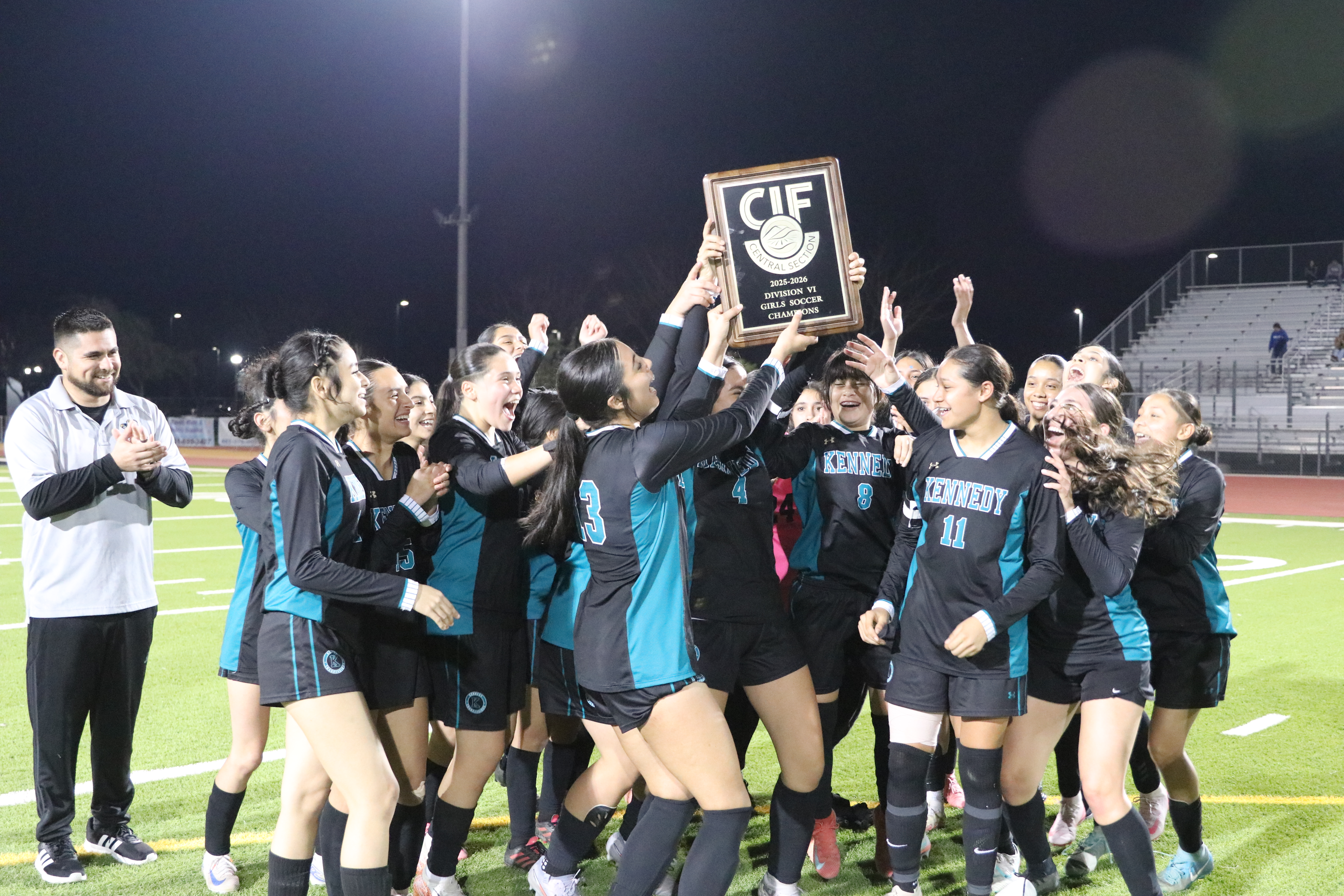 A group of young female athletes, wearing matching uniforms, are celebrating and holding up a trophy on a sports field at night, with a stadium and lighting visible in the background.