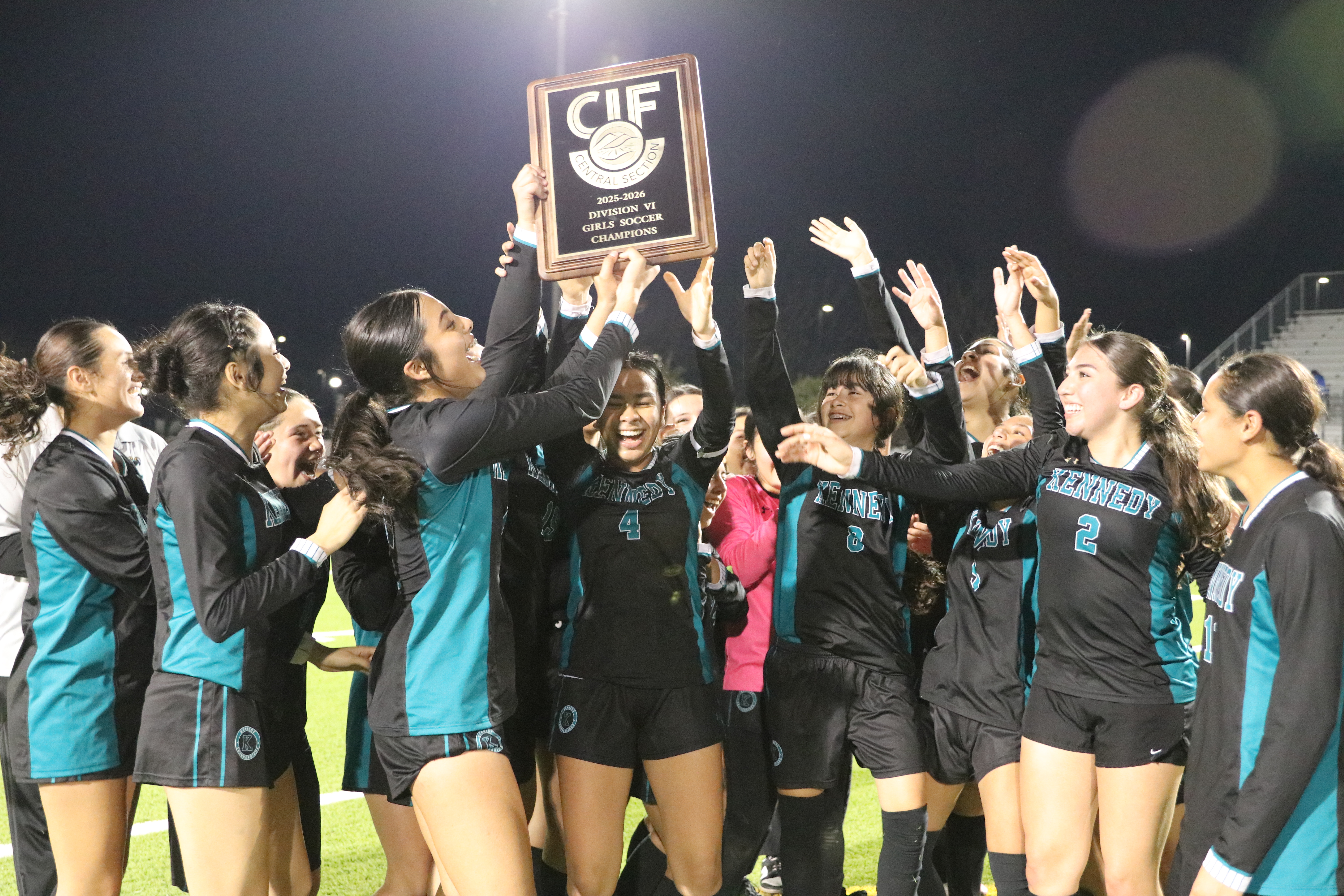 A group of young female athletes in sports uniforms are celebrating and holding up a trophy, with a dark night sky and stadium lights visible in the background.
