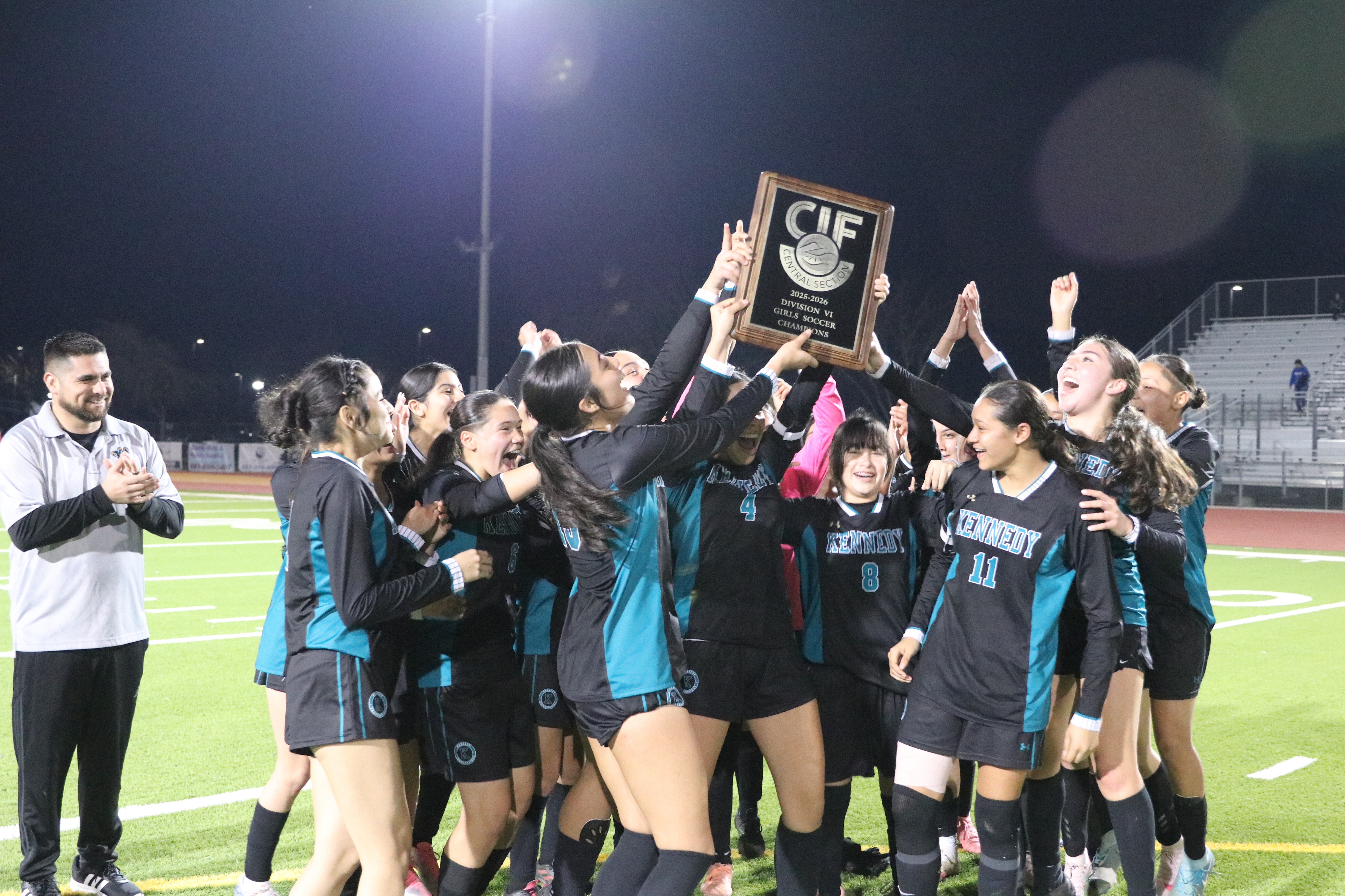 A group of young female soccer players celebrating their victory, holding up a trophy on a sports field at night.