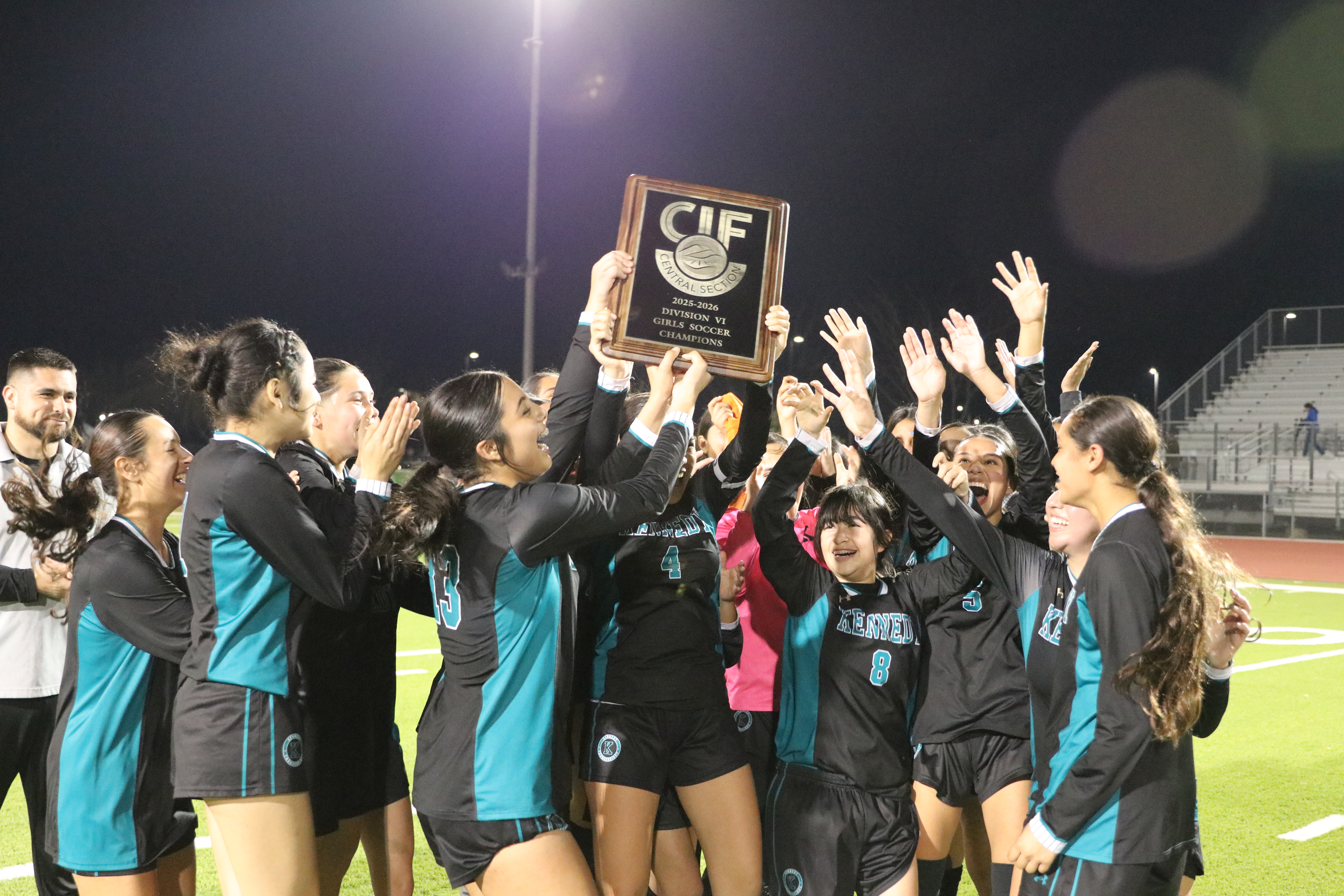 A group of people, likely a sports team, celebrating and holding up a trophy or award on a sports field at night.